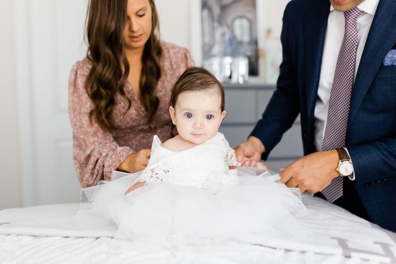 A small child in white sits on a bed surrounded by adults in formal wear.