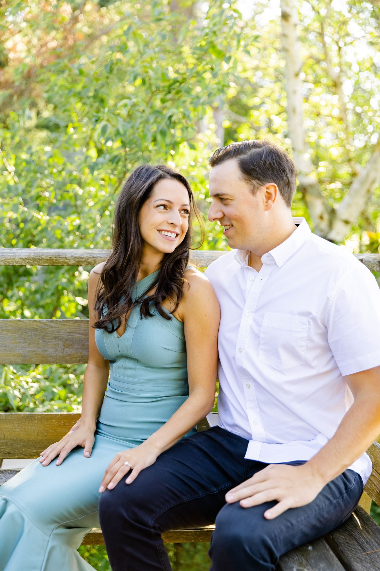 A couple shares an intimate moment while sitting on a wooden bench surrounded by lush greenery.