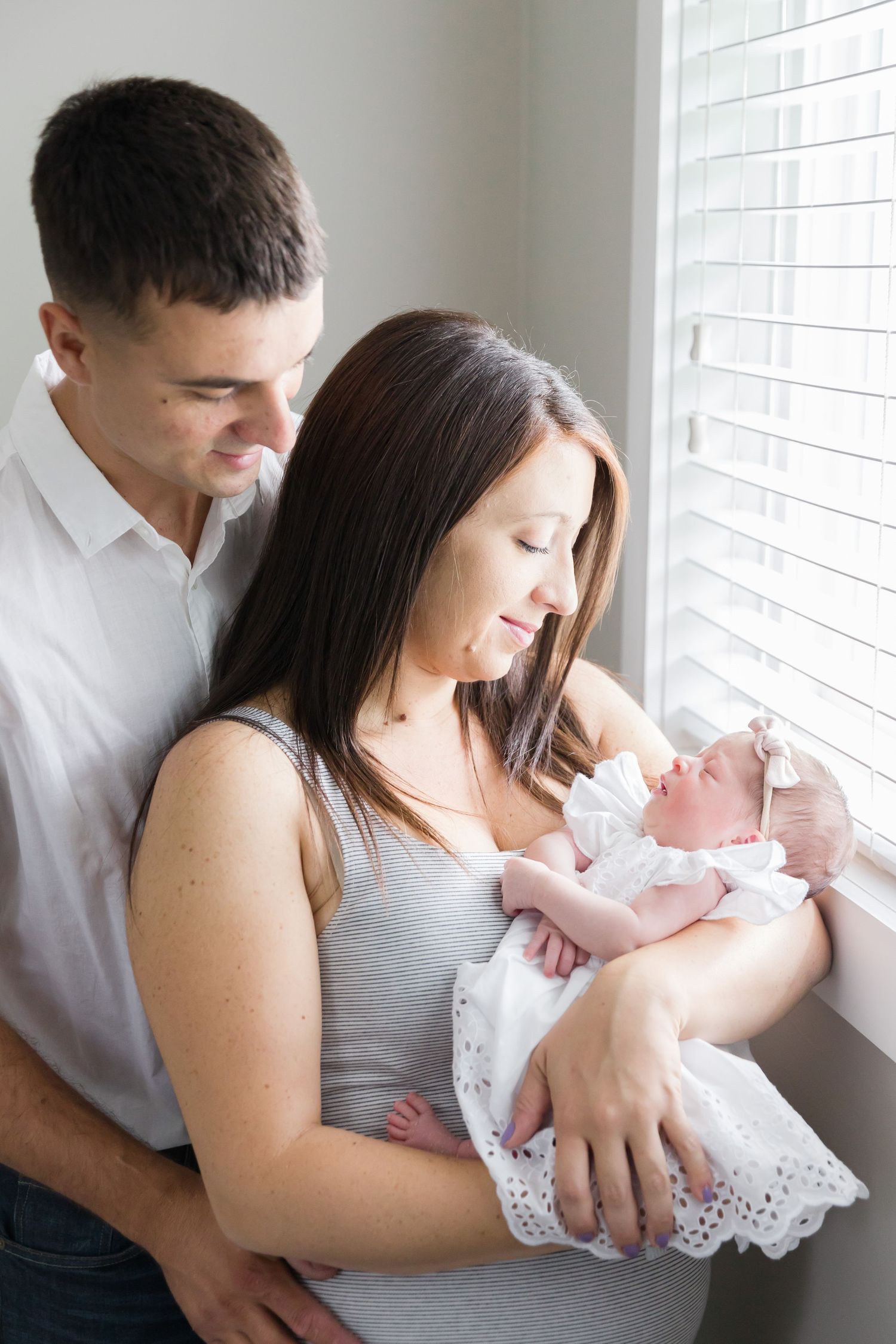 Parents by window lovingly gazing at baby in white dress.