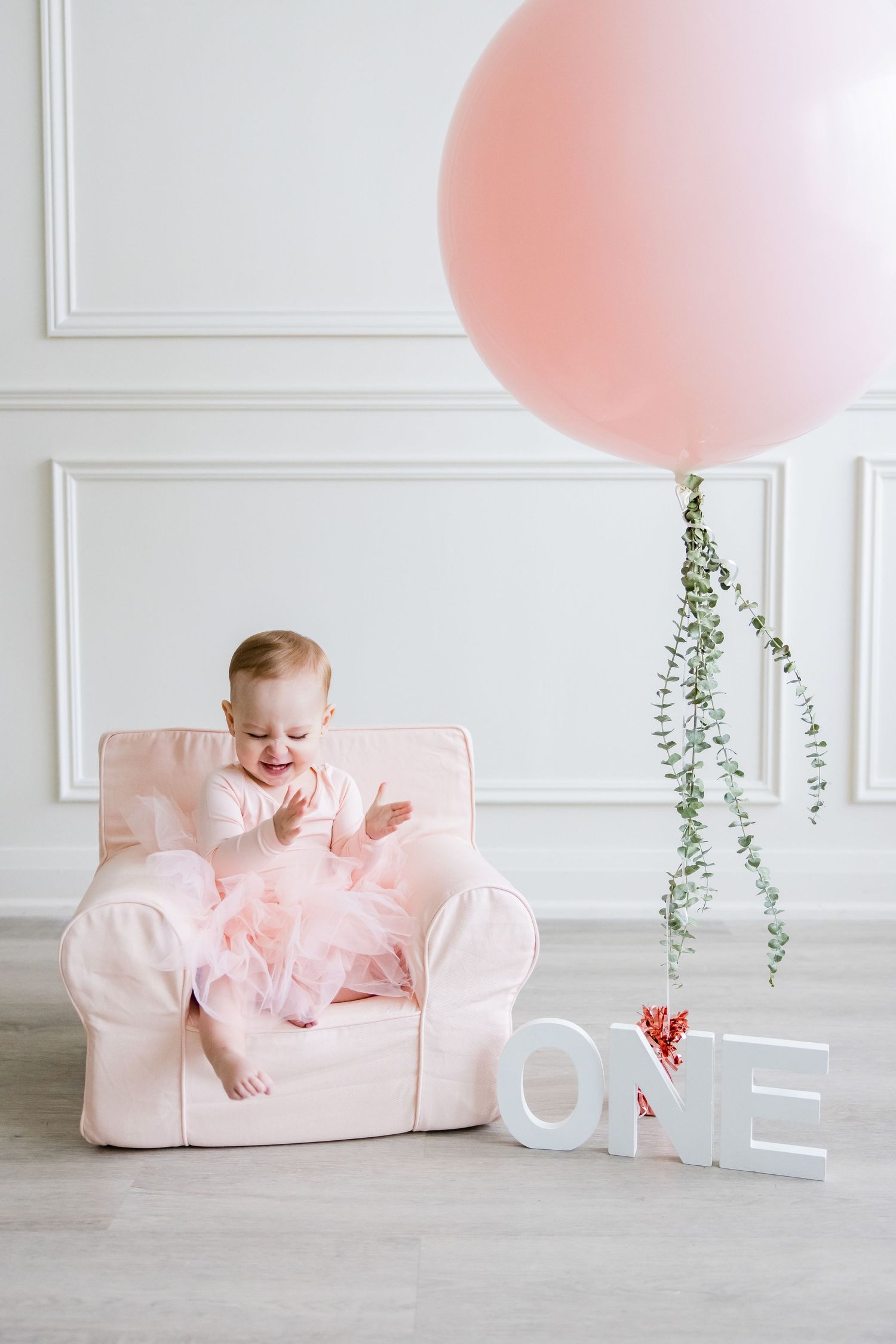 Pink first birthday photo with a large balloon, plush chair and white 'ONE' letters against white paneled wall.