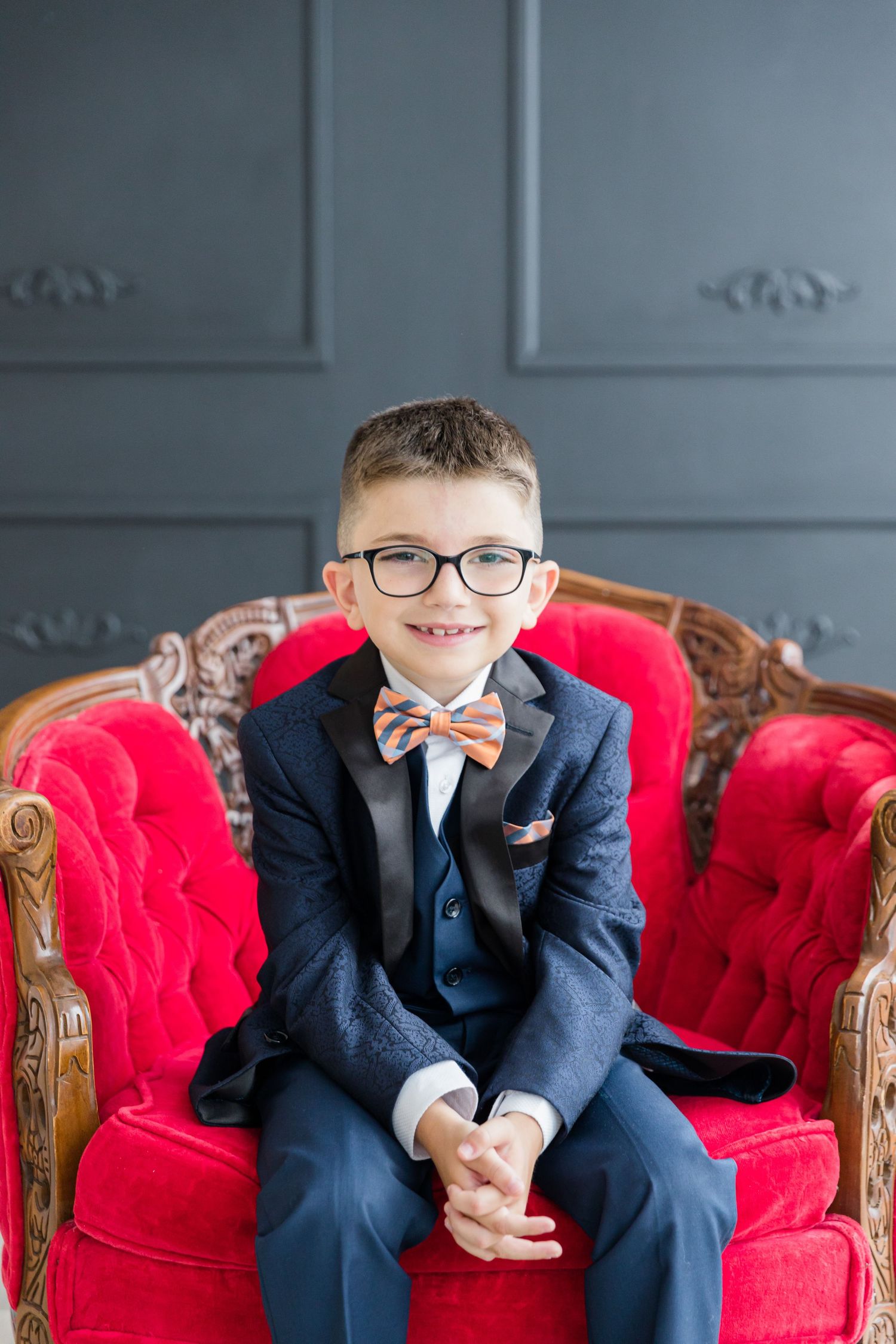 Child in navy suit and bow tie sits on ornate red velvet chair against gray wall.