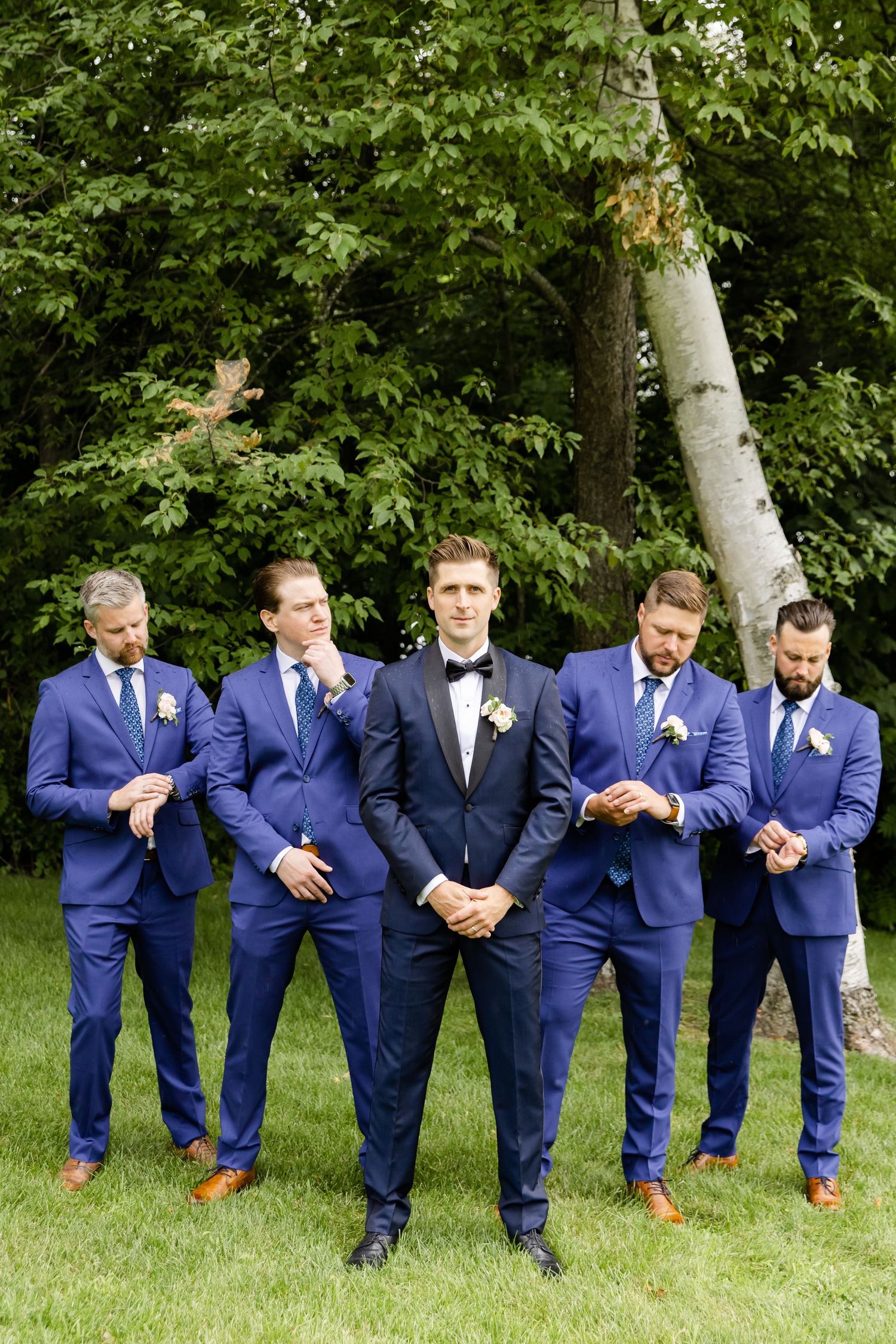 Groomsmen in navy suits and groom in black tux pose casually on green lawn.