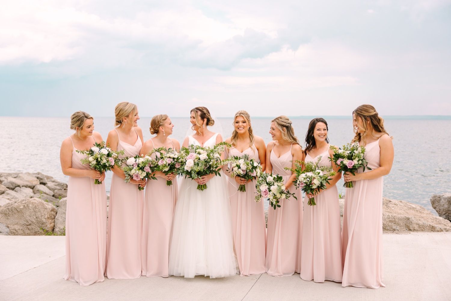 Bridesmaids and bride hold bouquets while lined up against an oceanfront backdrop.