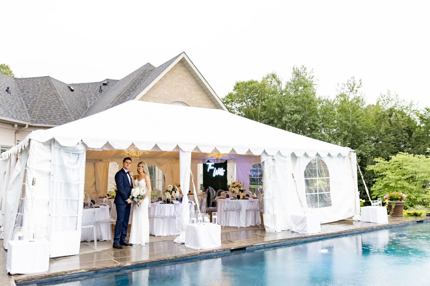 A reception tent set up next to a swimming pool with white tables and chairs inside.