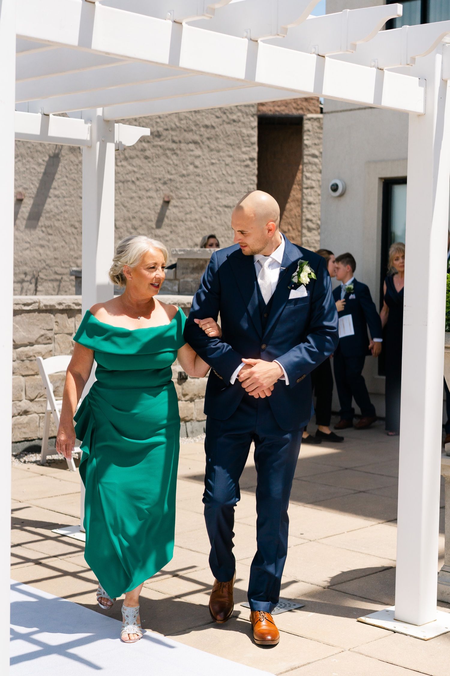A pair walks together under a white pergola, with one wearing emerald green and the other in navy.