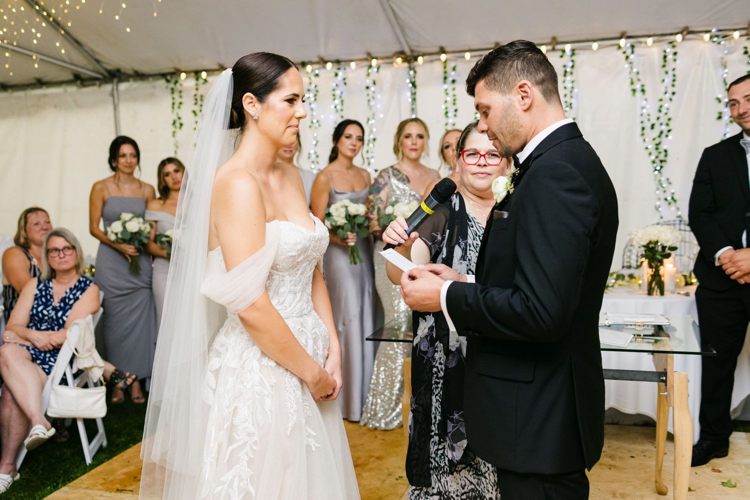 Wedding vow exchange in front of guests under tent with string lights and greenery.