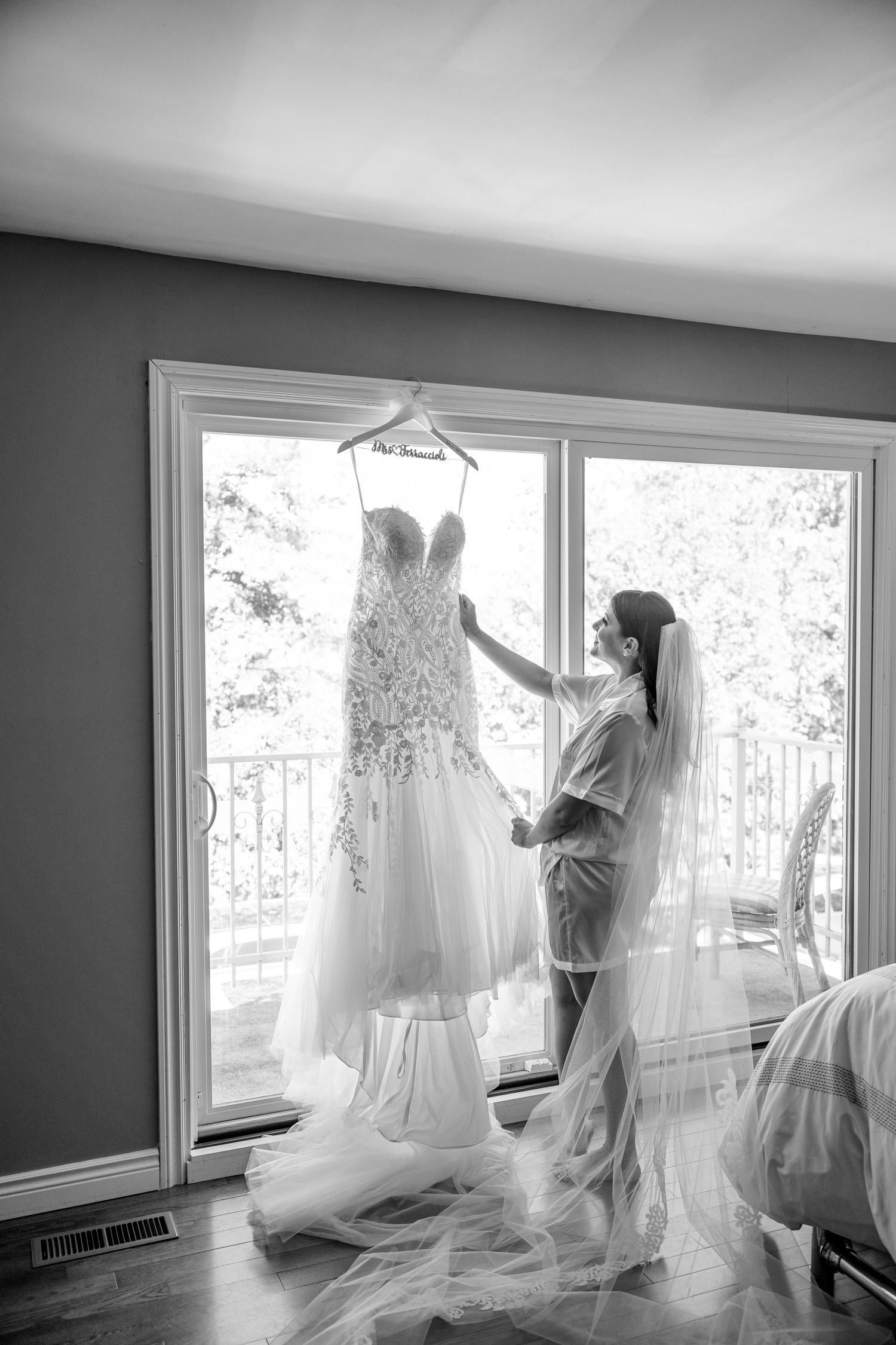 A flowing white lace wedding dress displayed in front of large windows with natural backlight streaming in.
