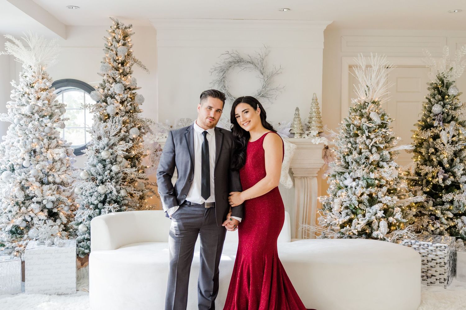 Elegantly dressed couple pose together in front of decorated white Christmas trees in a bright interior setting.