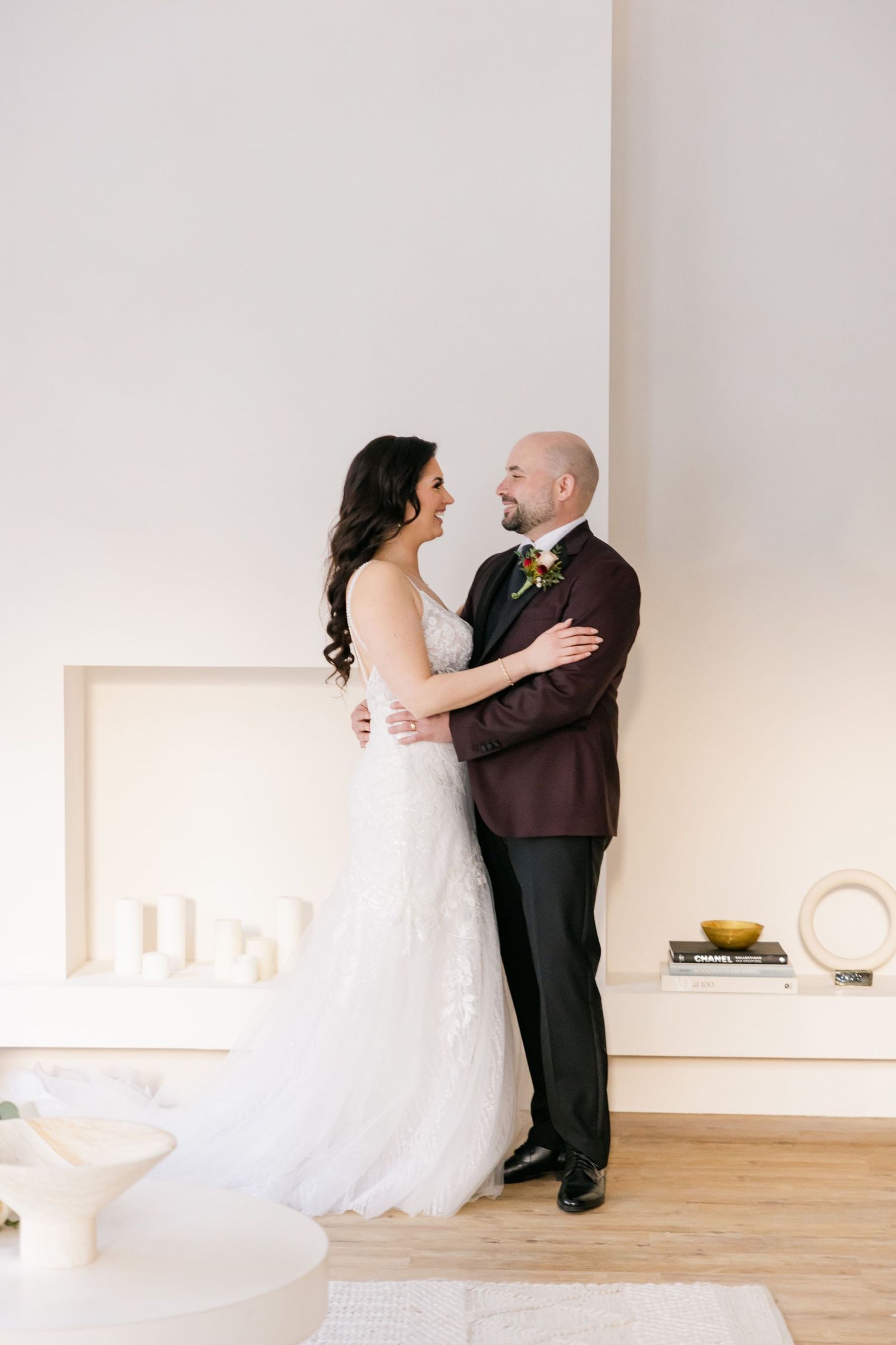 Newlyweds embrace in an elegantly decorated white room with hardwood floors.