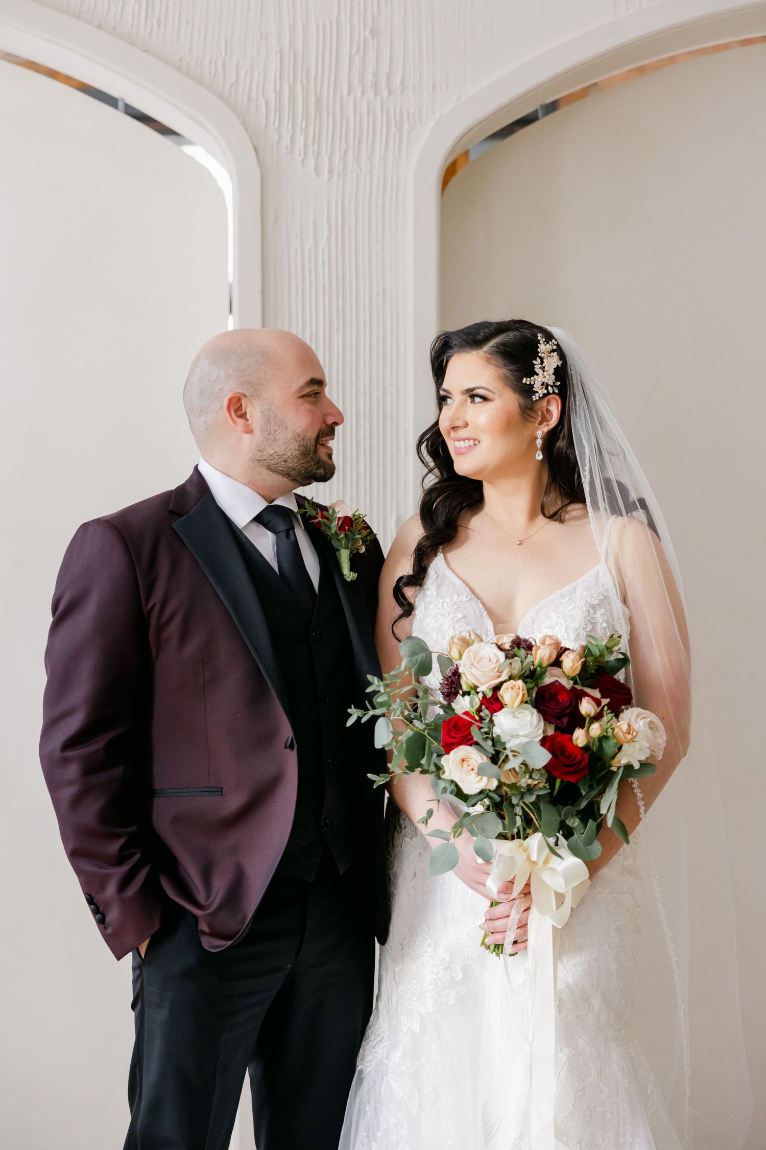 Wedding couple posing under arched doorway with burgundy and white floral bouquet.