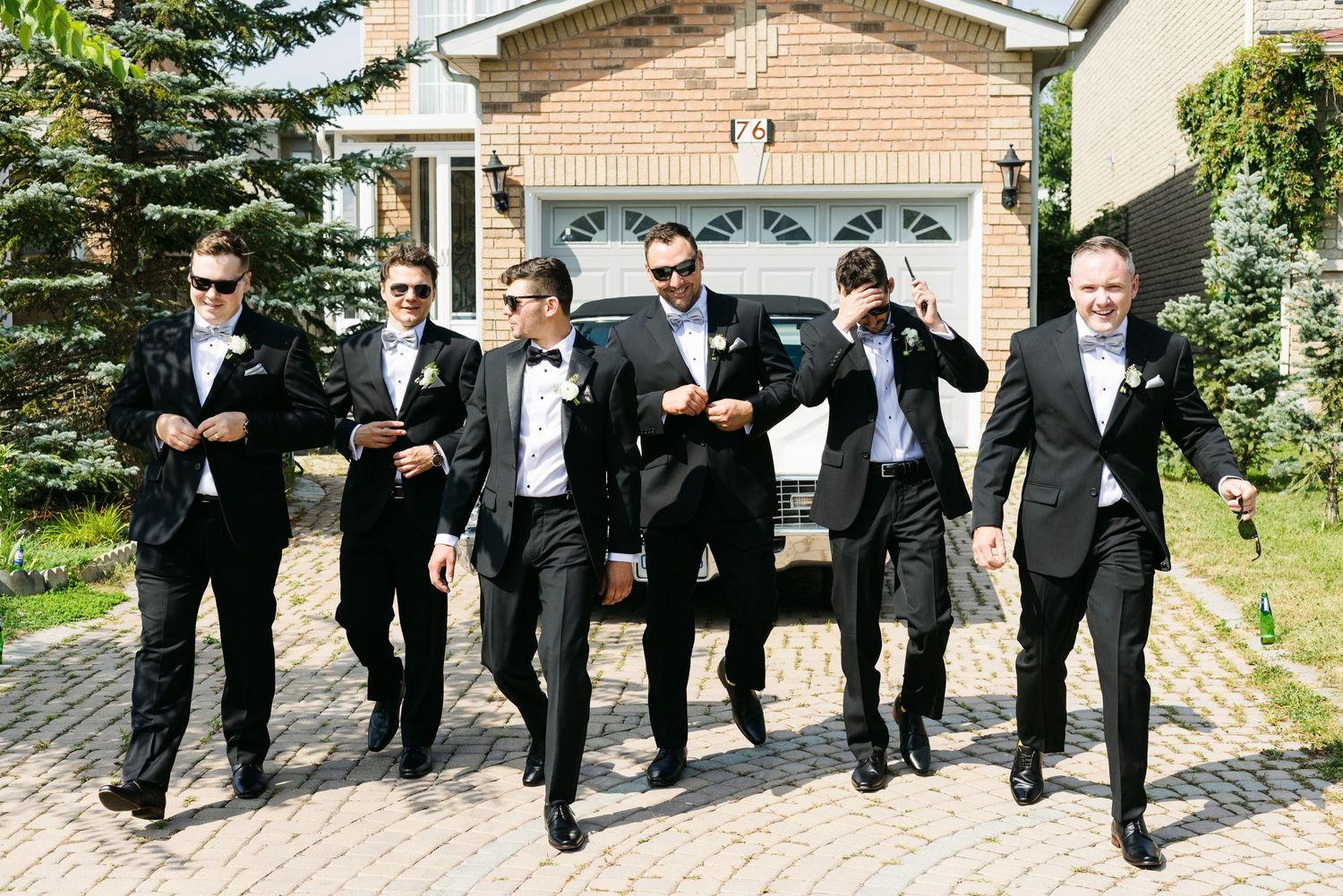 Groomsmen in black tuxedos striking dynamic poses on brick driveway.