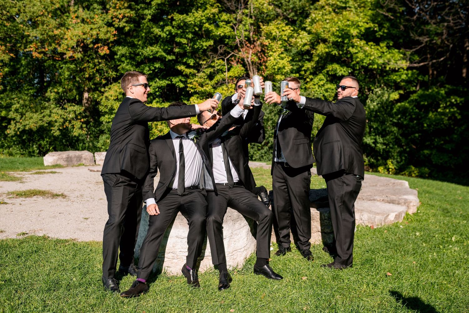 Groomsmen in black suits pose playfully for a group photo outdoors.