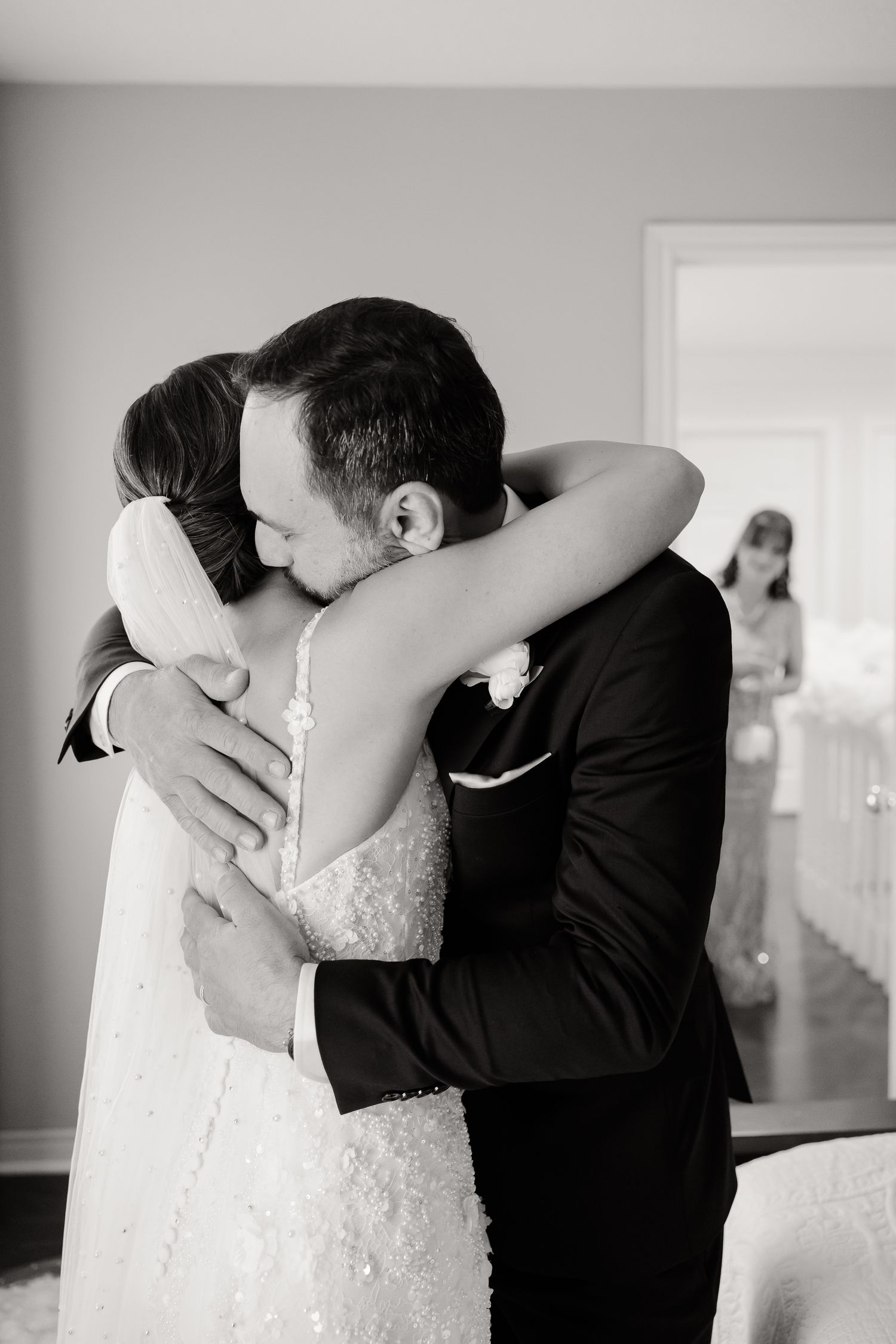 A tender black and white wedding moment shows a couple embracing while another figure stands in the background.