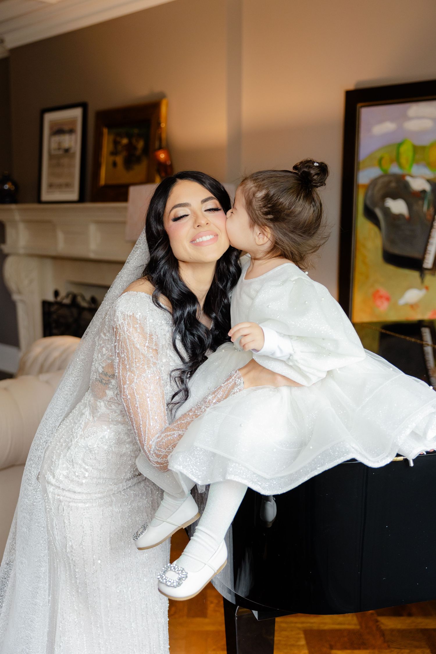 A sweet moment is captured between a bride and young attendant by a piano during wedding preparations.