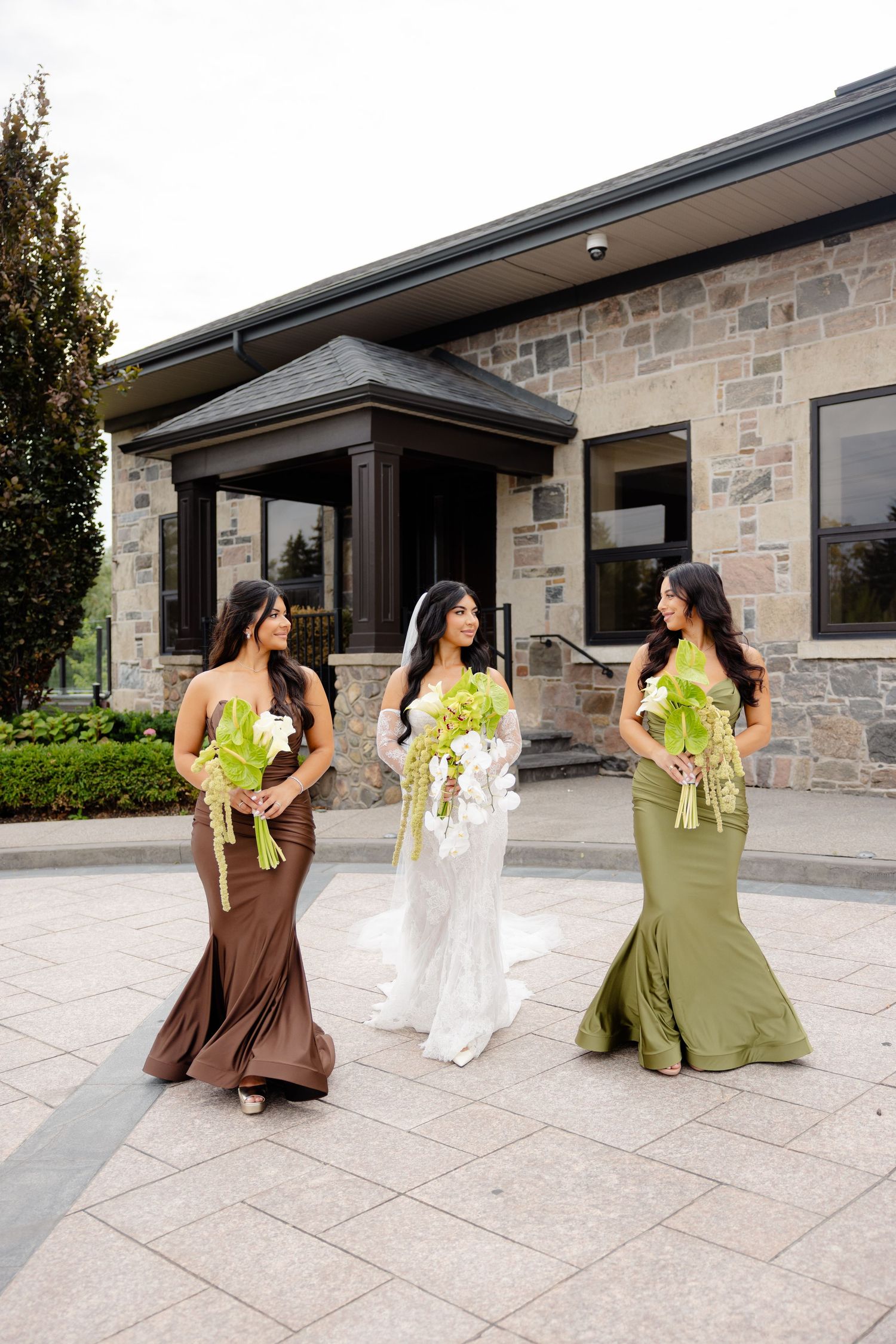 Bridesmaids in brown and olive green dresses pose playfully on a stone patio during a wedding celebration.