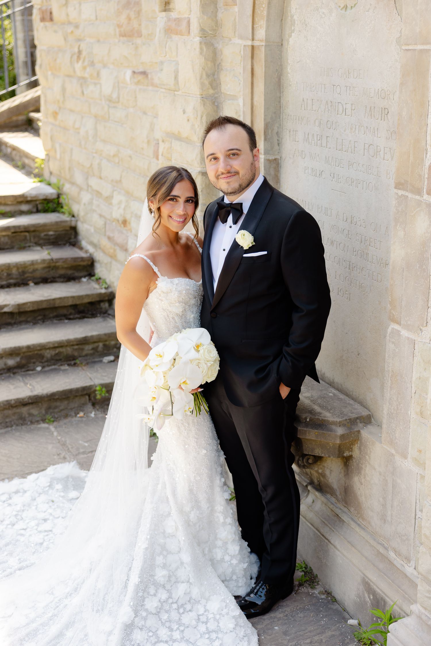 A couple on their wedding day posing together on stone steps with the bride holding a white floral bouquet.
