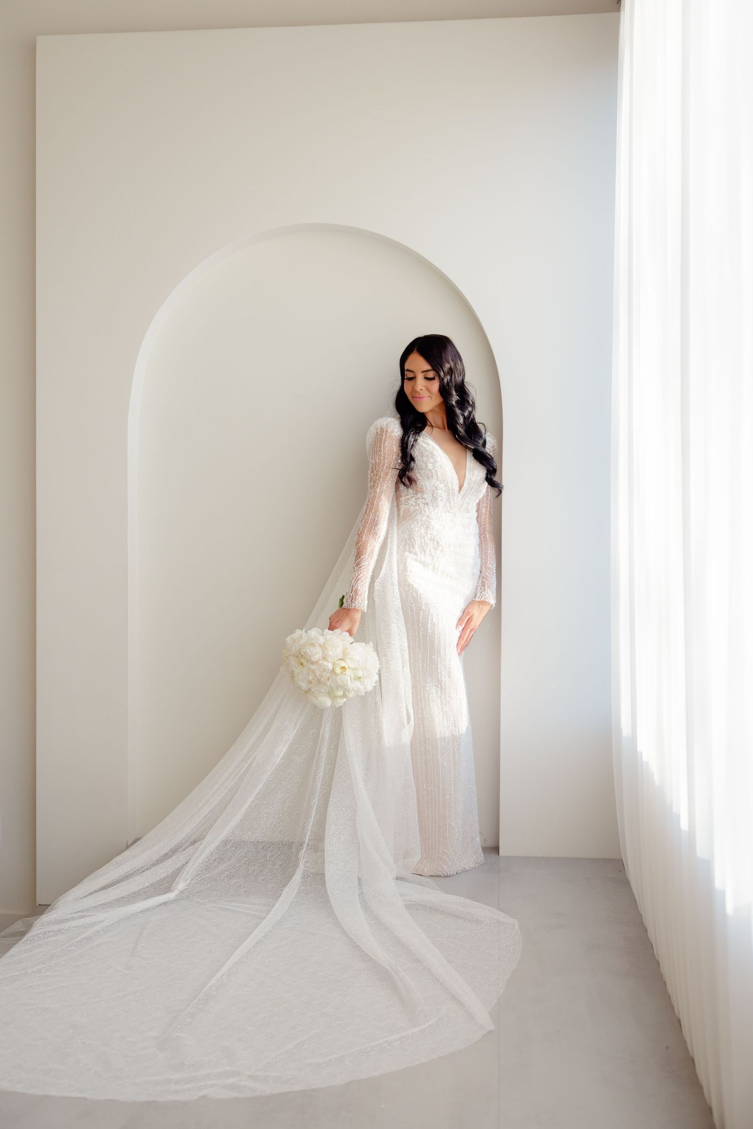 Elegant bride in long sleeve lace wedding gown with cathedral veil poses in minimalist white archway.