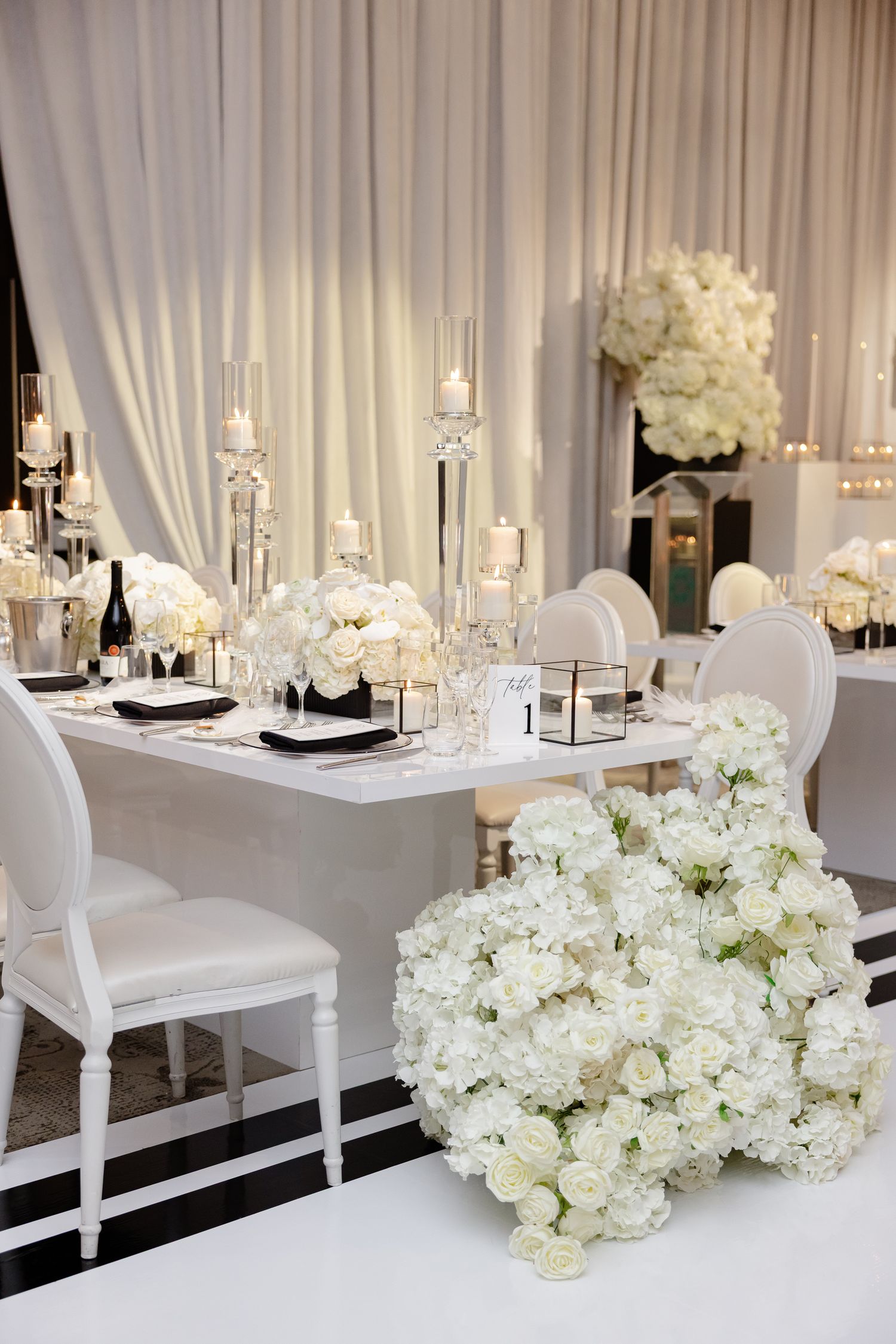 Elegant white and silver wedding reception table with tall crystal candle holders, white roses, and modern ghost chairs.
