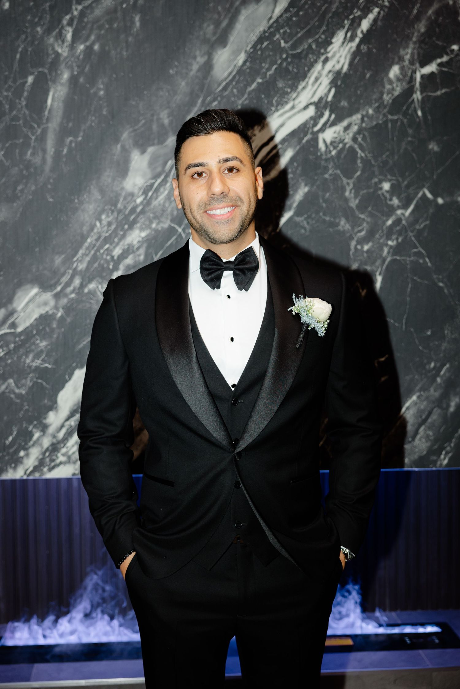 Dapper groom in three-piece tuxedo with boutonniere stands before marble backdrop.