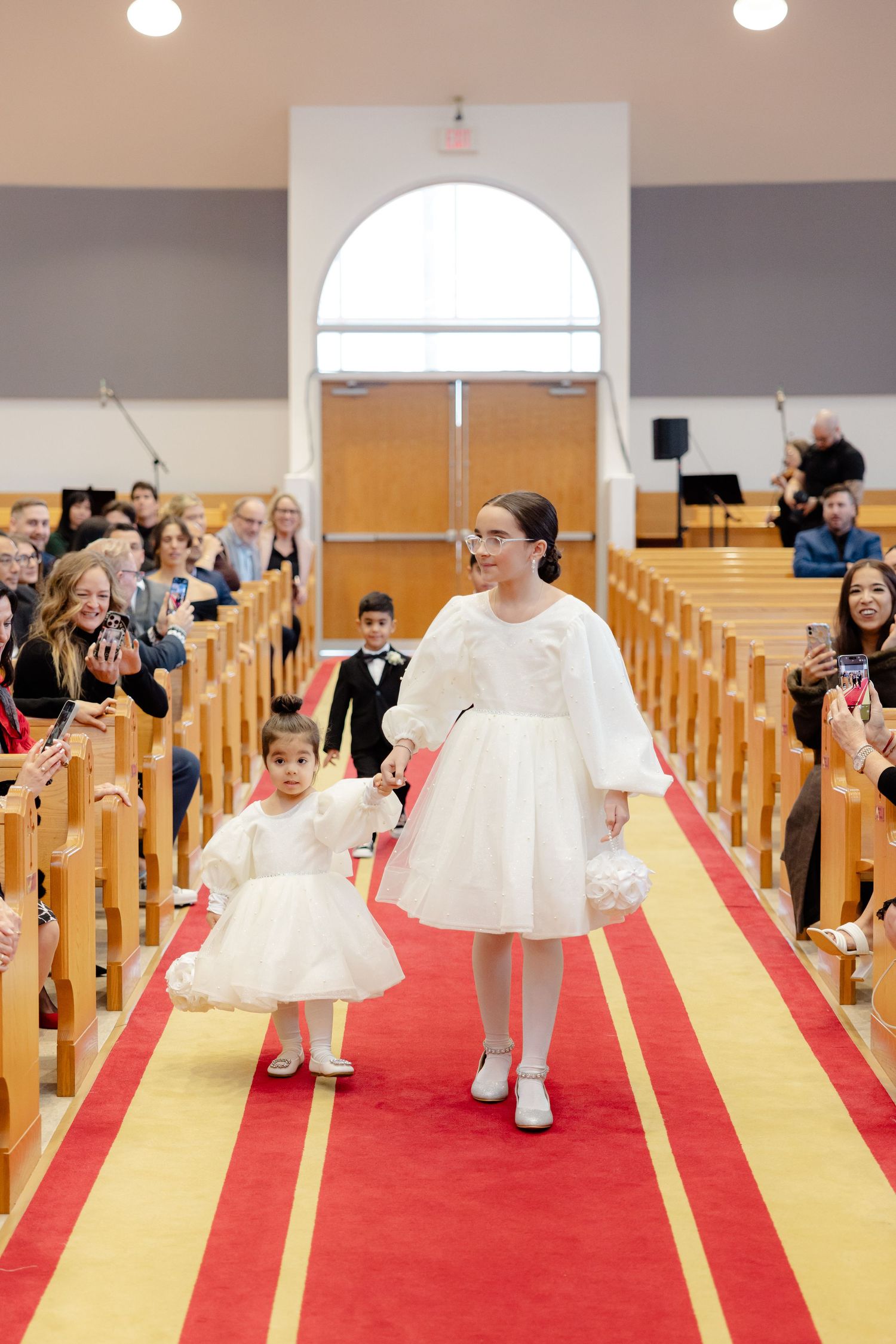 Two figures in white dresses walk down a red and gold church aisle while guests watch from pews.