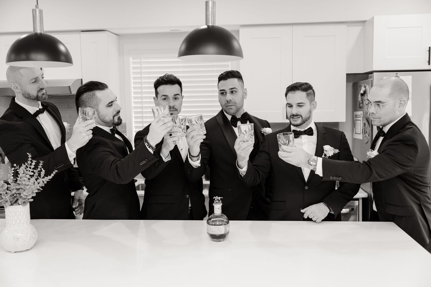 Black and white photo of wedding party toasting with drinks at kitchen counter.