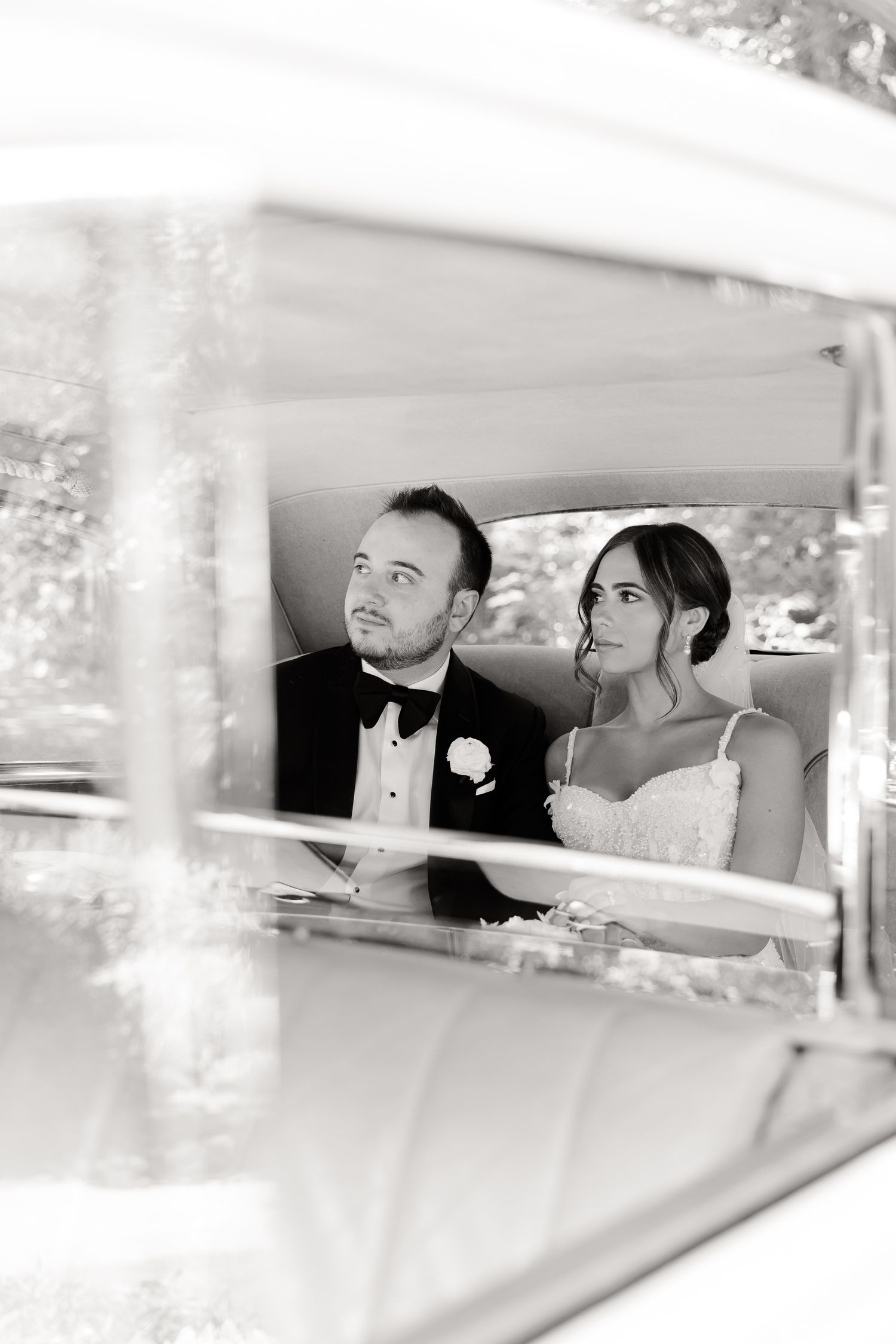 Black and white photo of a couple sitting in a vintage car on their wedding day smiling through window.