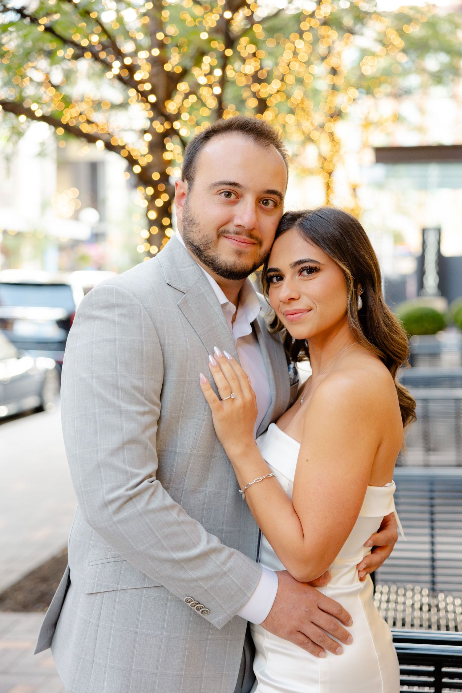 An engaged couple poses in front of twinkling lights, with the woman displaying her ring while wearing a white dress.