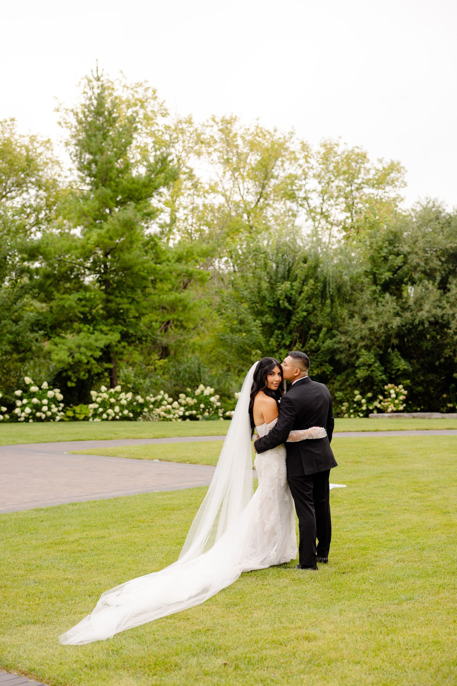 A couple in wedding attire share a romantic moment on a lush green lawn with blooming trees in the background.