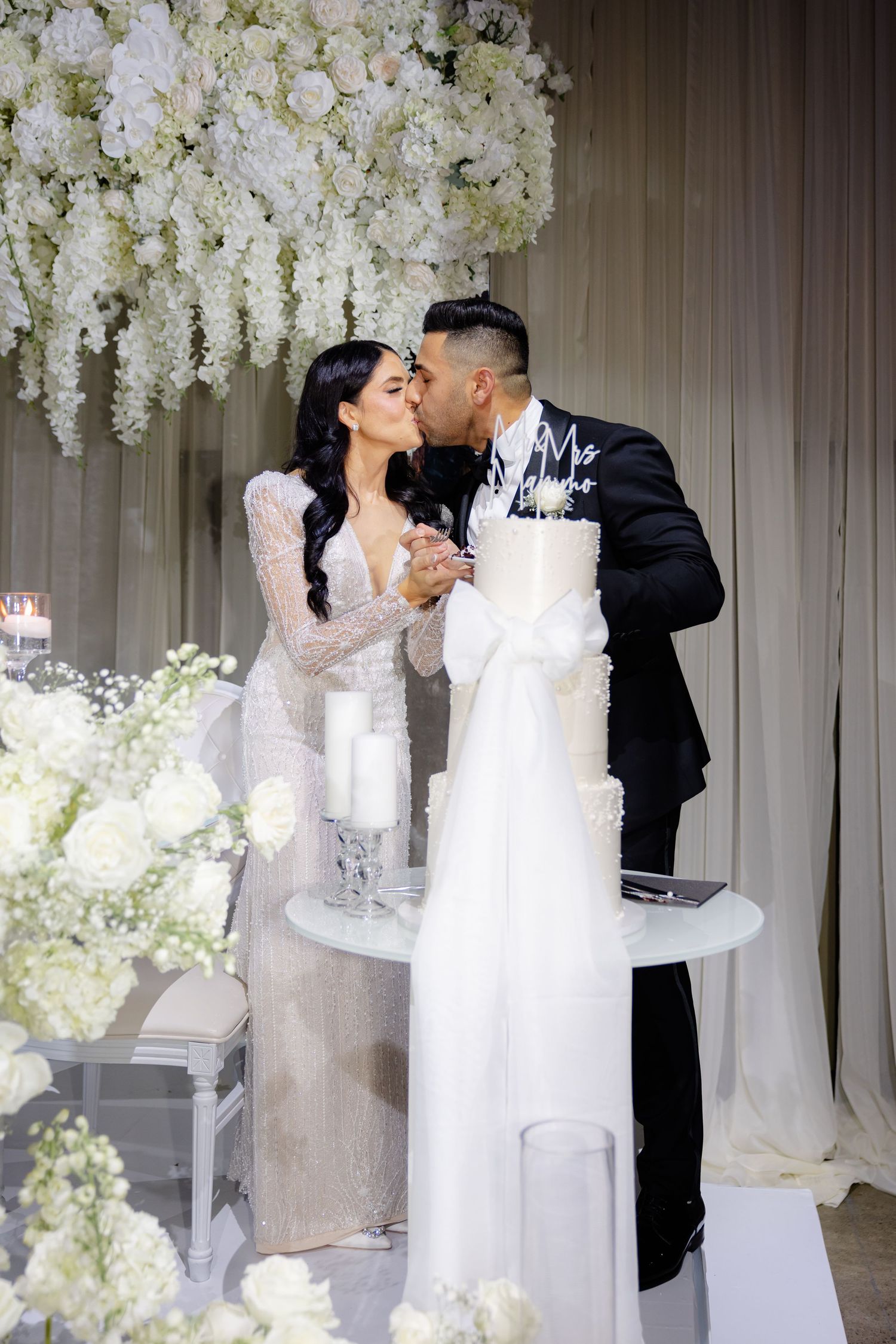 Newlyweds share intimate moment while cutting cake under cascading white flowers.