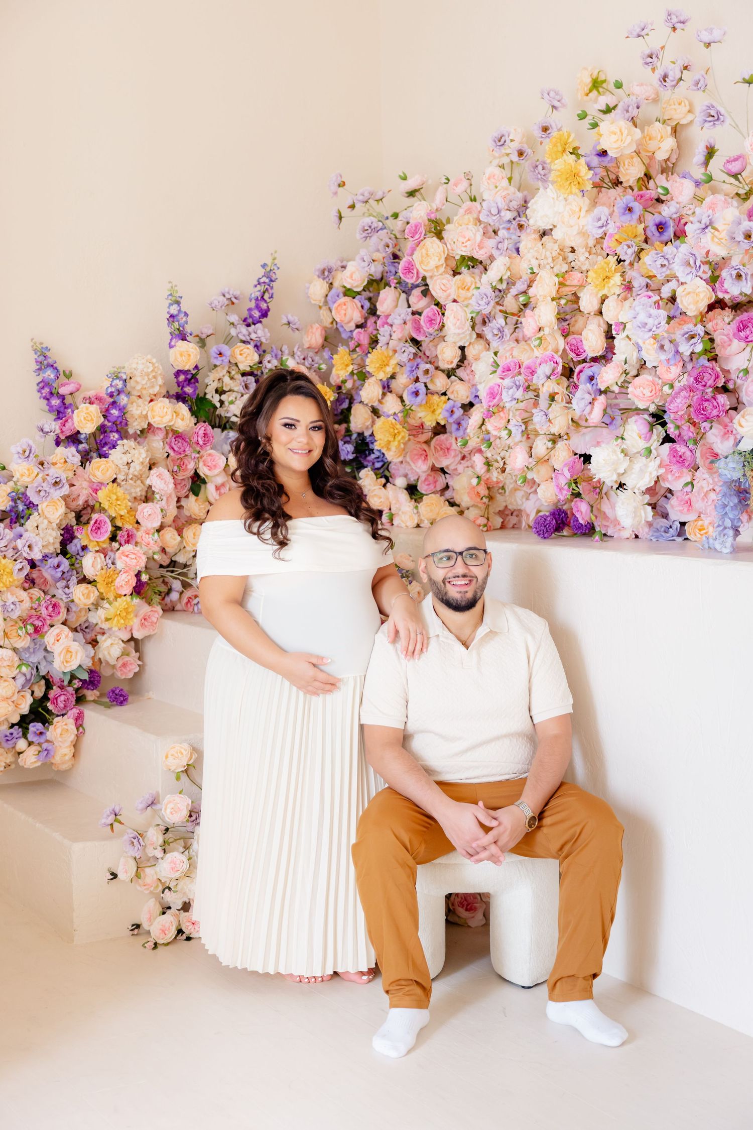A couple poses against a stunning floral wall backdrop with pink, purple and yellow blooms in a bright studio setting.