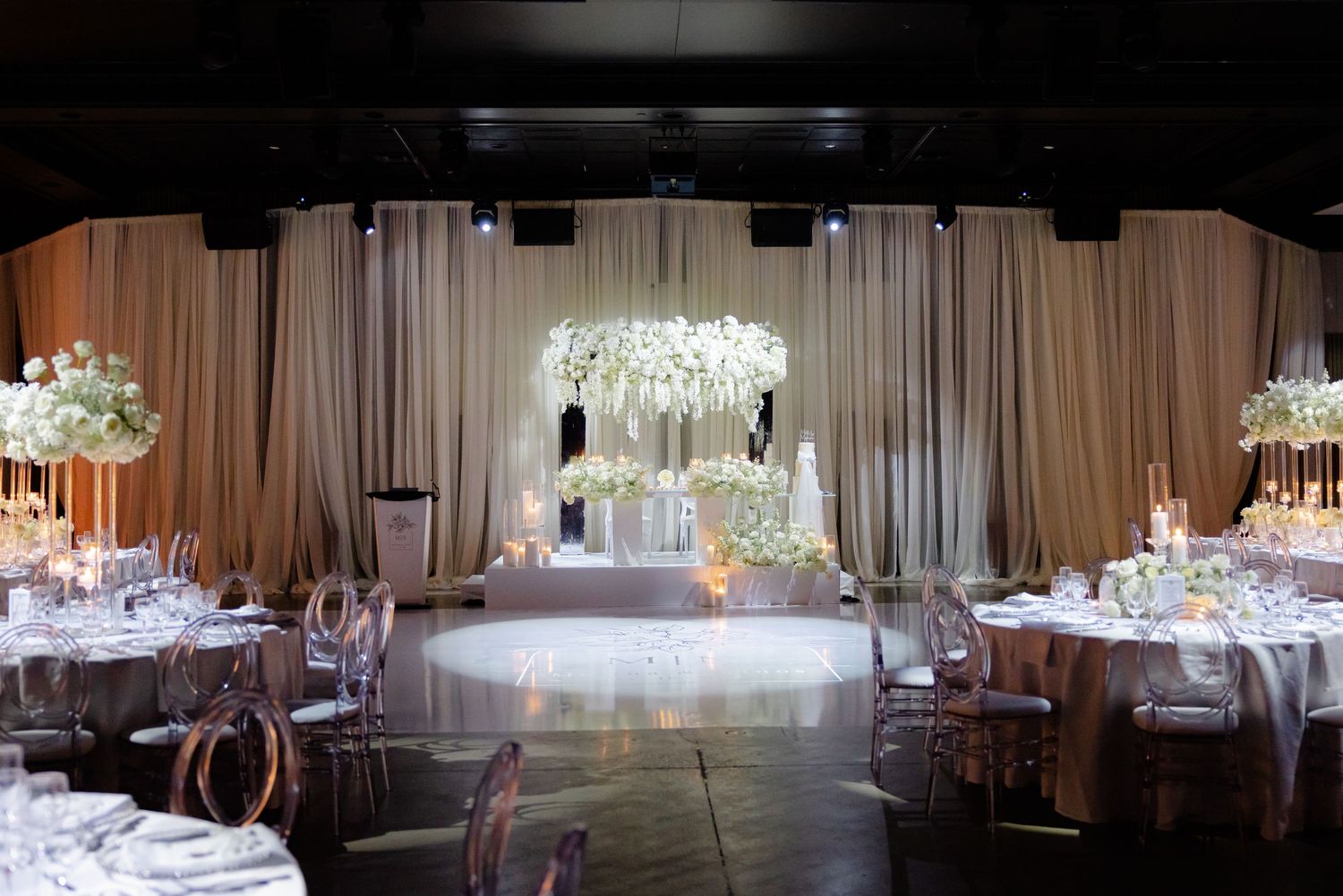 Wedding reception venue with white draping and illuminated floral arch at the focal point.