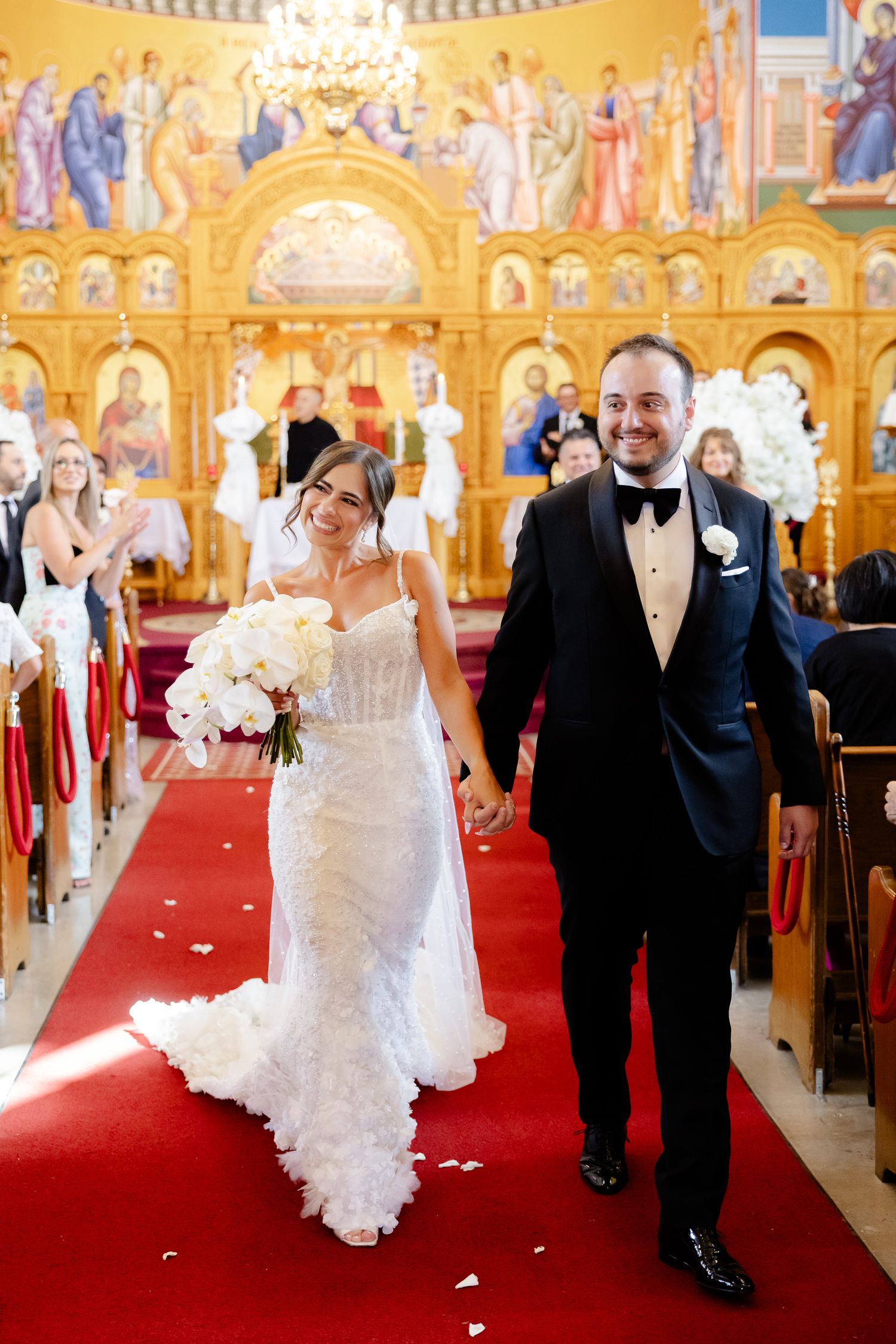 A wedding couple walks down a red-carpeted church aisle with ornate golden religious murals adorning the walls.