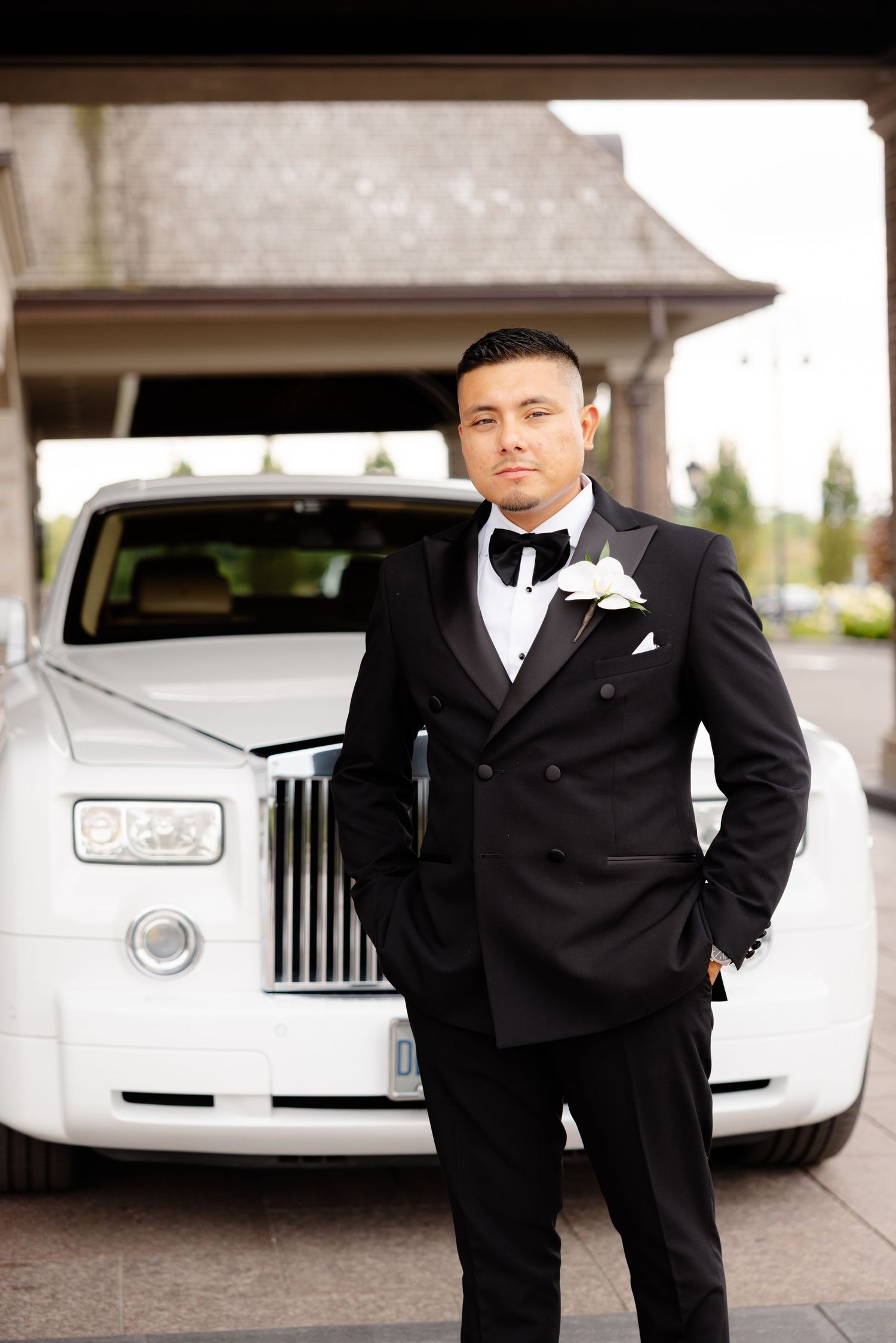 A man in a classic black tuxedo poses confidently next to a white Rolls Royce luxury car.