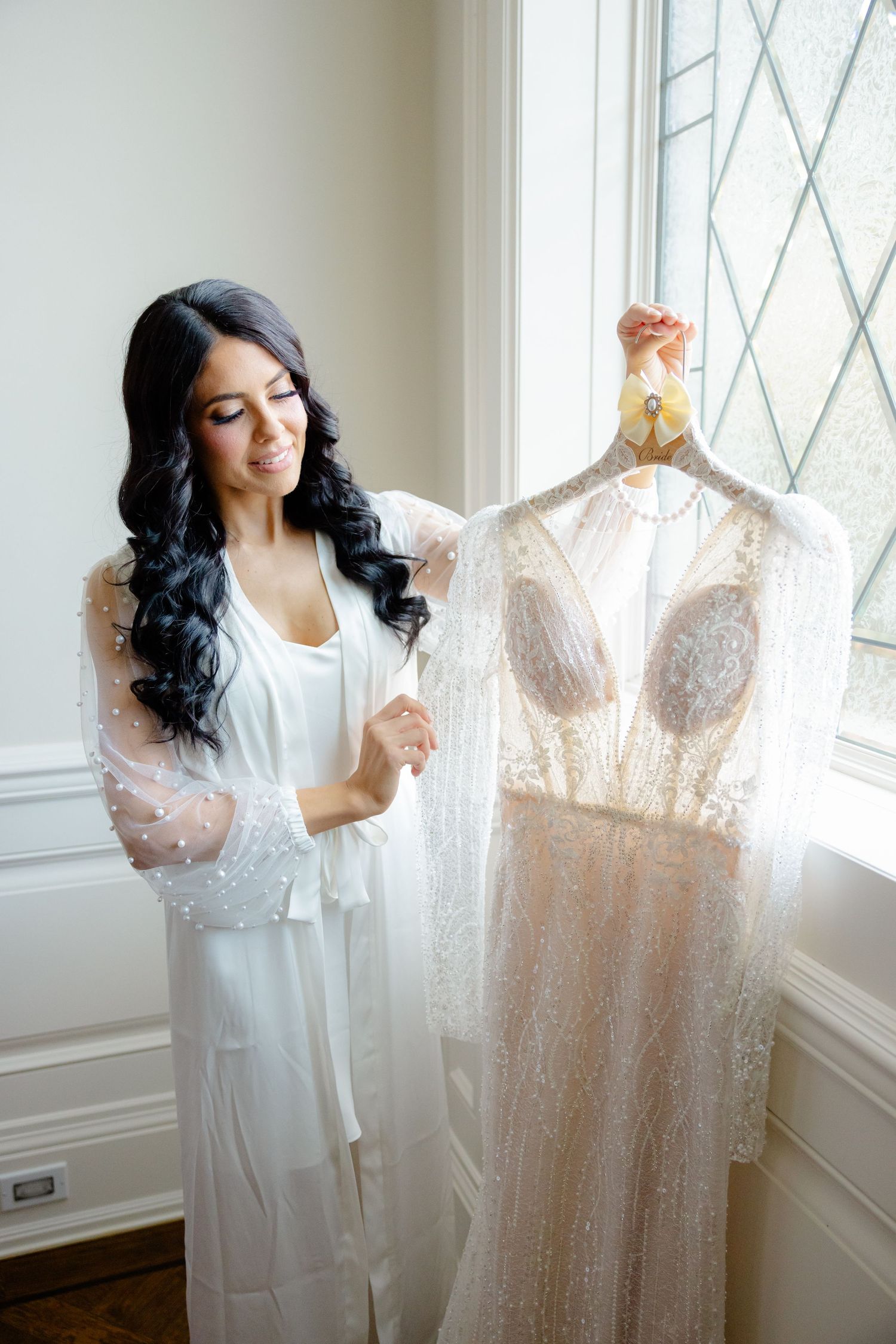 Woman in white robe examining delicate lace wedding gown hanging by window.