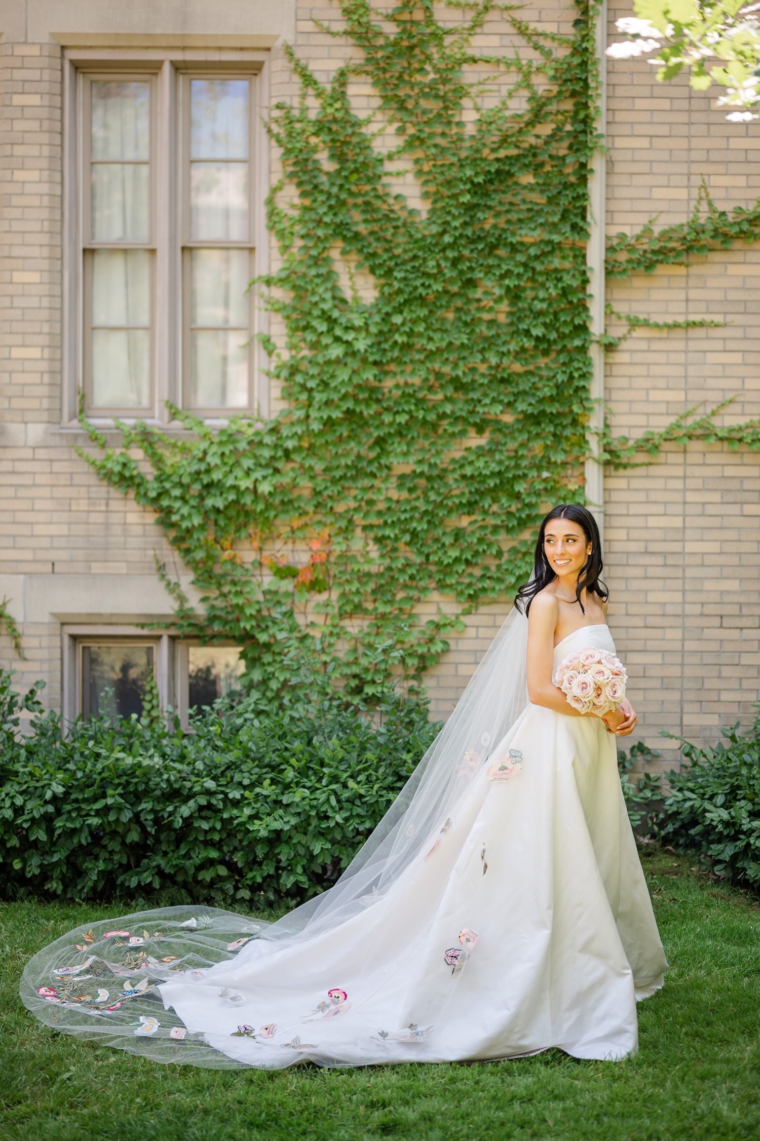 A bride in a white gown with floral embellishments poses against an ivy-covered building on a sunny day.