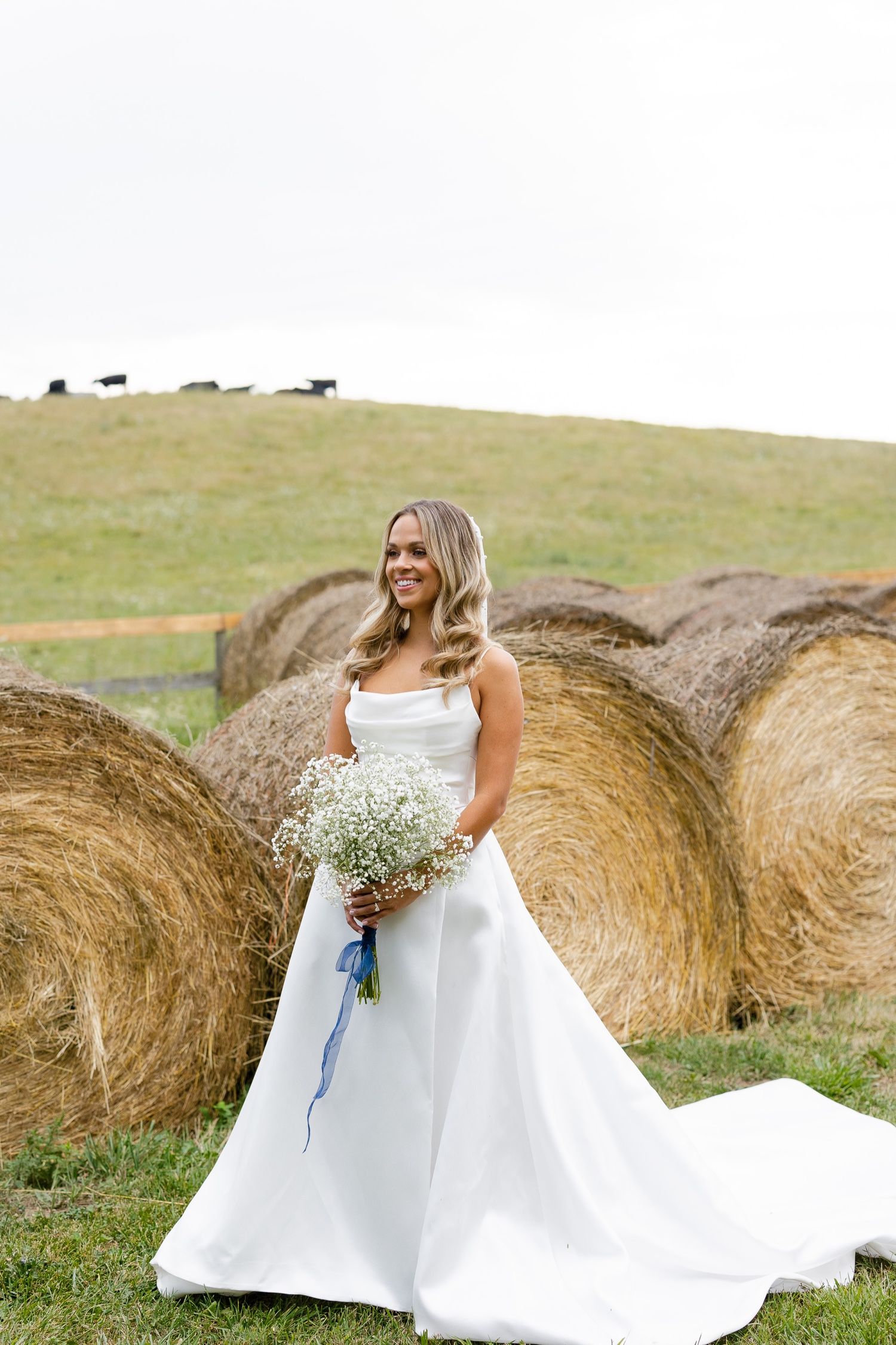 A bride in a white dress holds a baby's breath bouquet while standing in front of hay bales on a hillside.