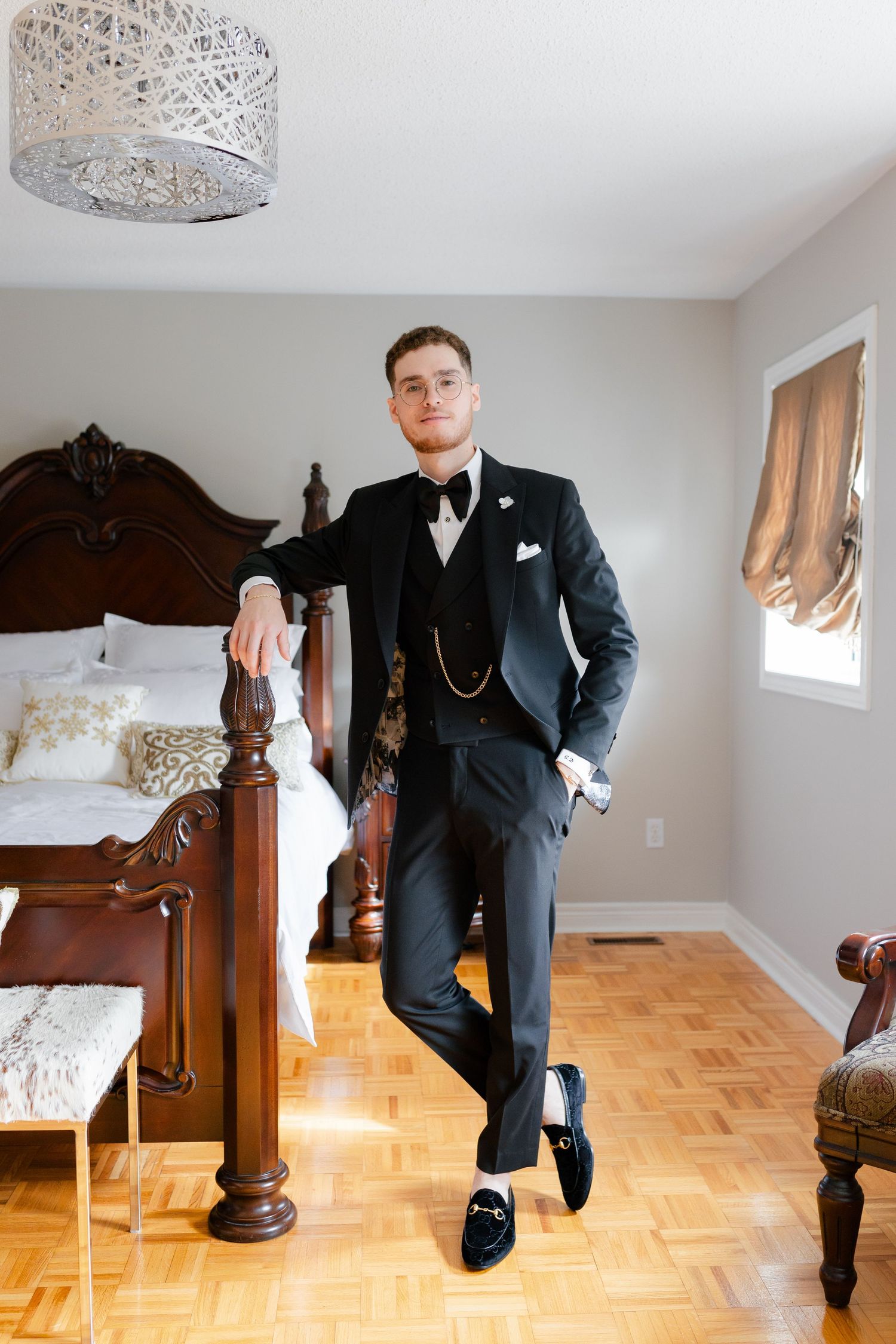 A formal portrait against a wooden bed frame showing a classic black tuxedo with pocket watch chain.