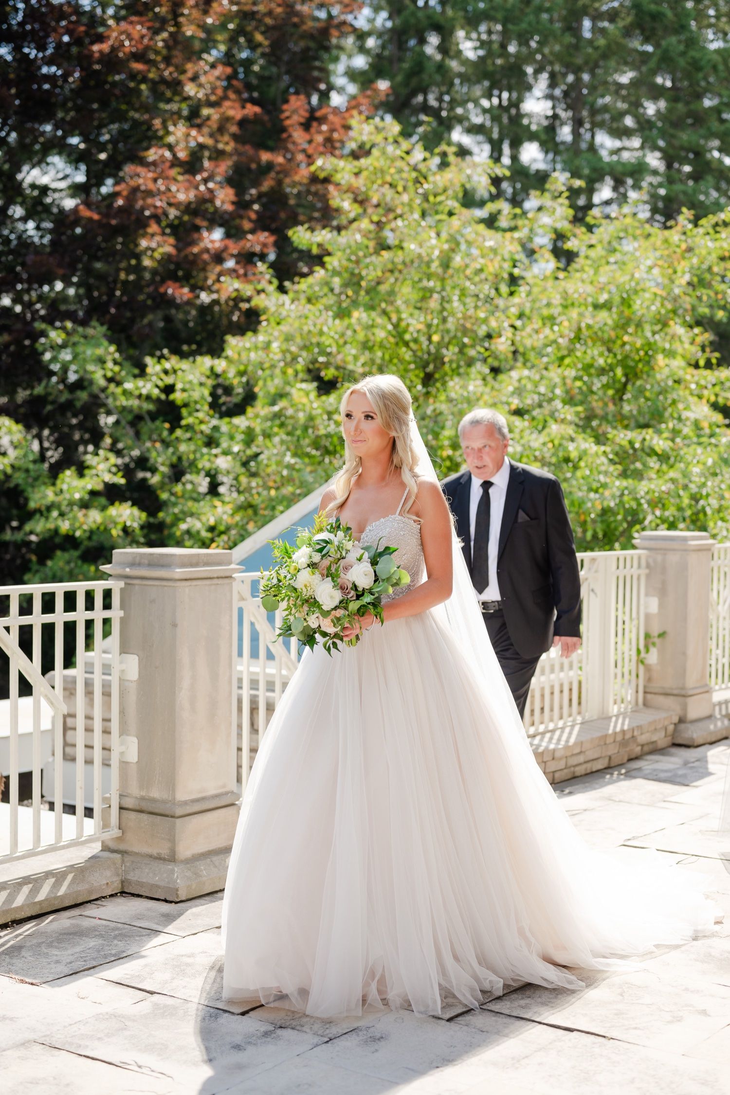 A bride in a flowing white ballgown holds a lush bouquet while walking on a stone terrace with garden backdrop.