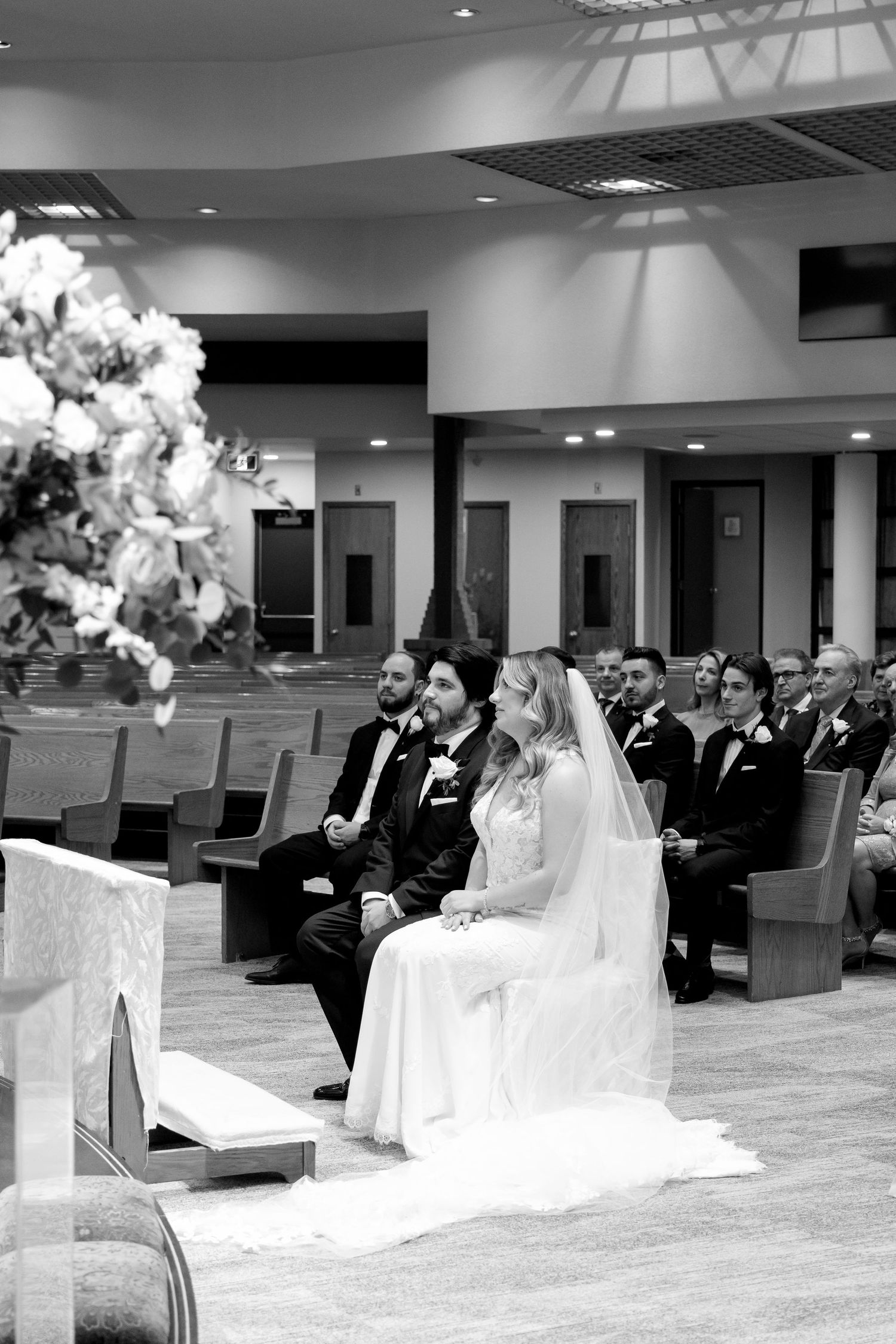 Black and white photo of wedding ceremony with couple seated in modern church setting.