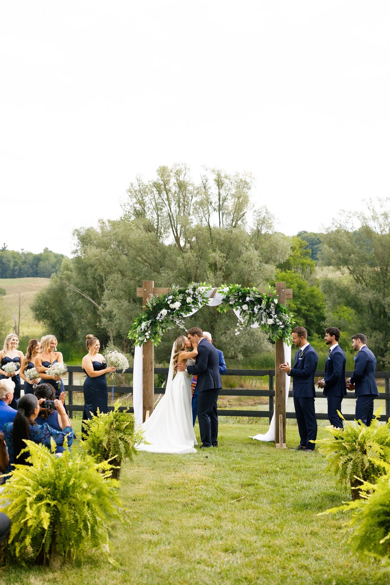 A wedding ceremony takes place under a floral arch with ferns lining the aisle in a scenic outdoor setting.