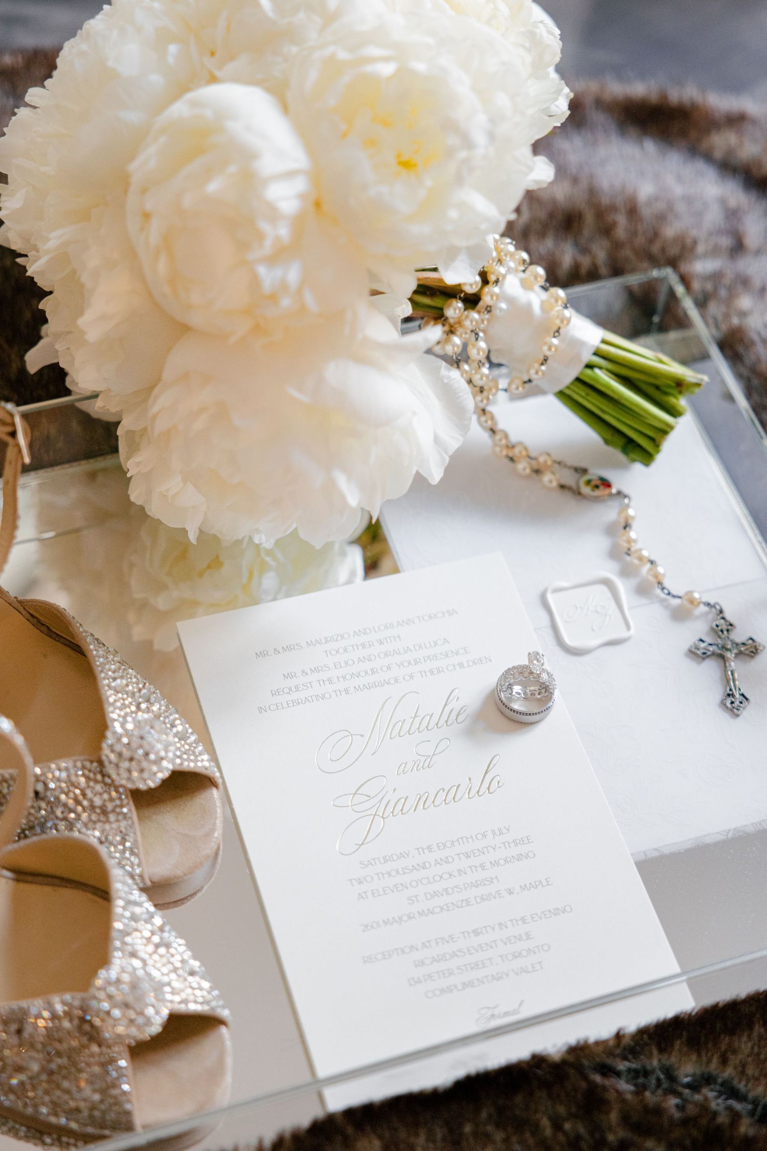 White peony bouquet with rosary beads and elegant wedding invitation on textured surface.