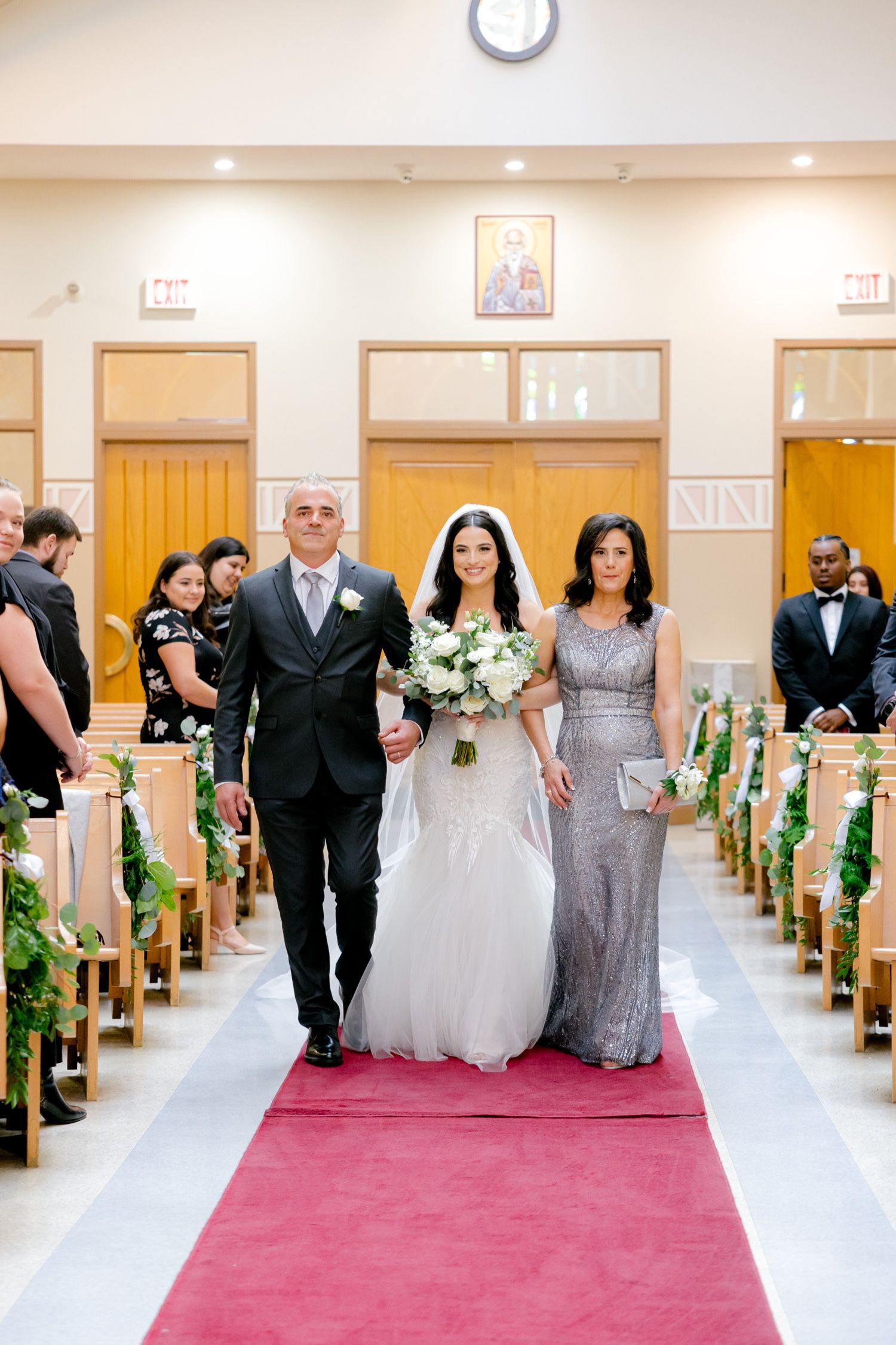 Wedding party walks down the aisle of a church decorated with greenery and a red carpet runner.