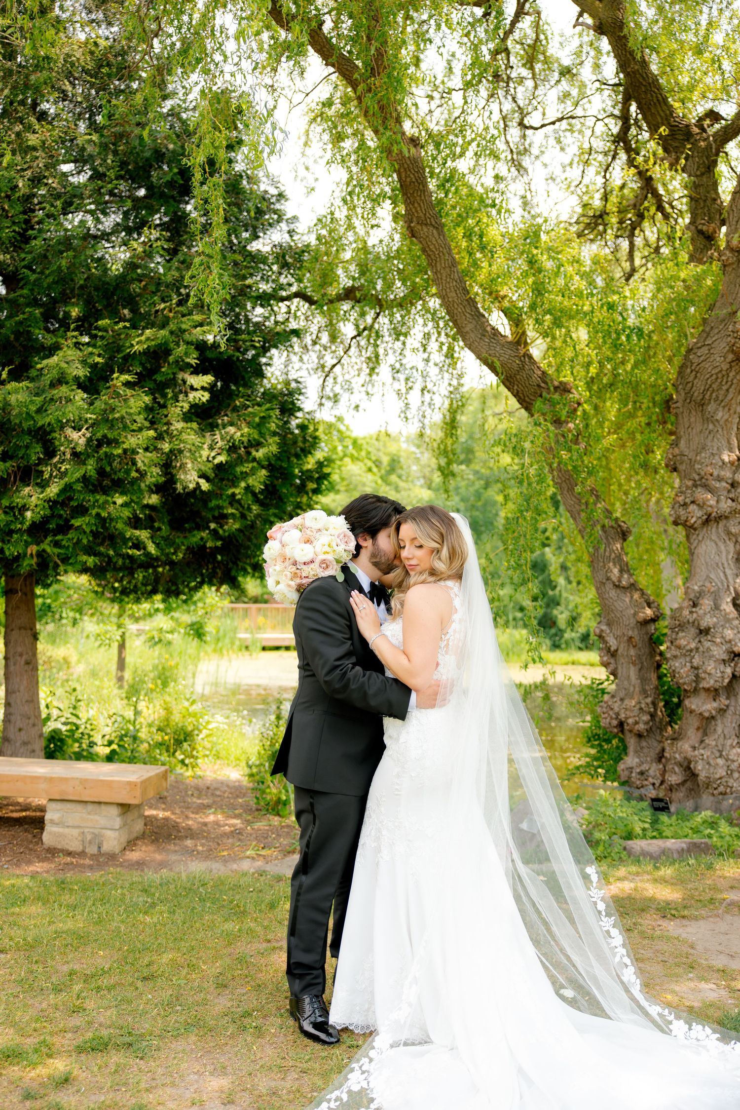 Wedding portrait under a majestic willow tree with flowing veil and pink floral bouquet.