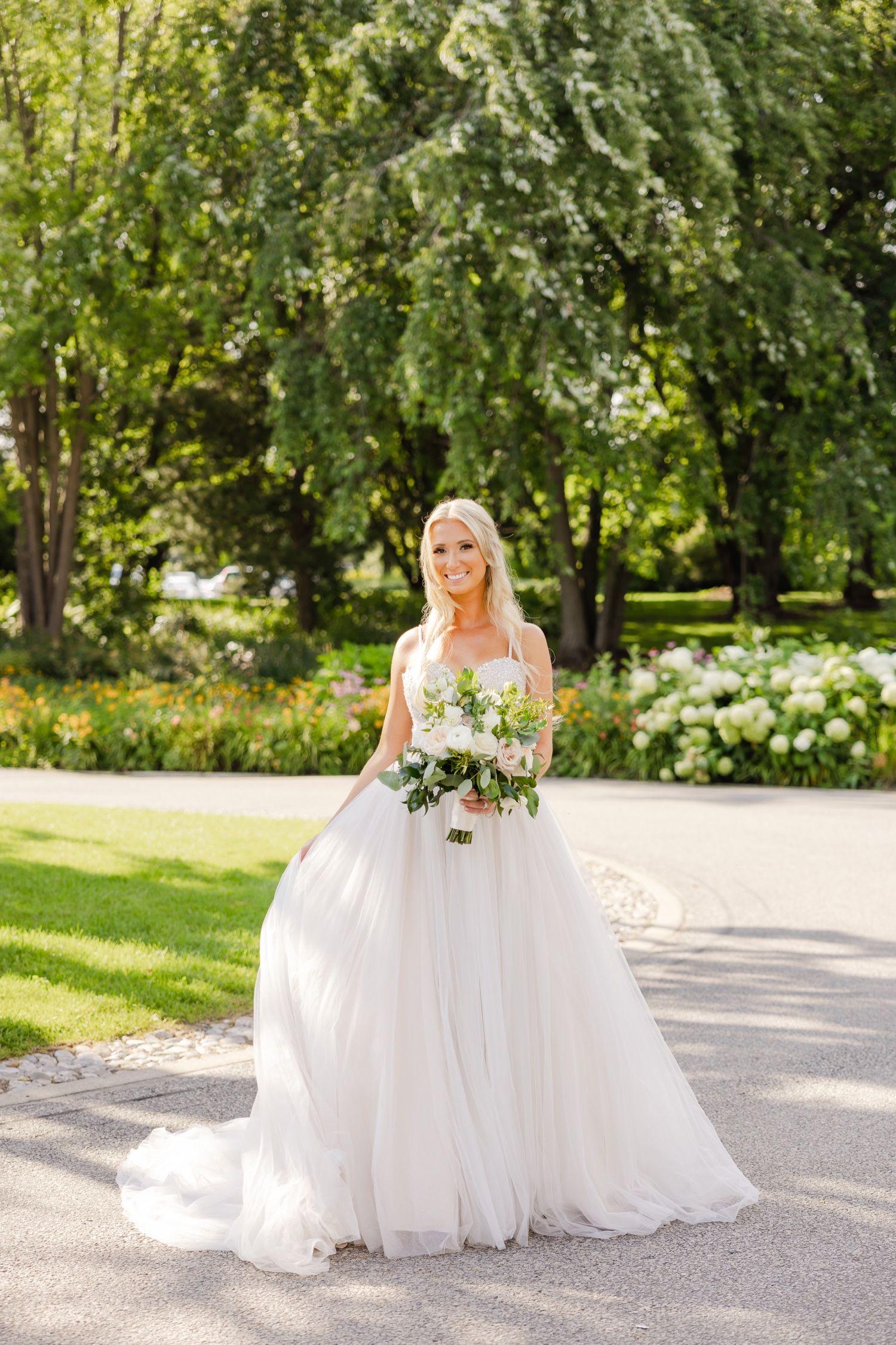 A bride in a white tulle gown poses on a garden pathway surrounded by green trees.