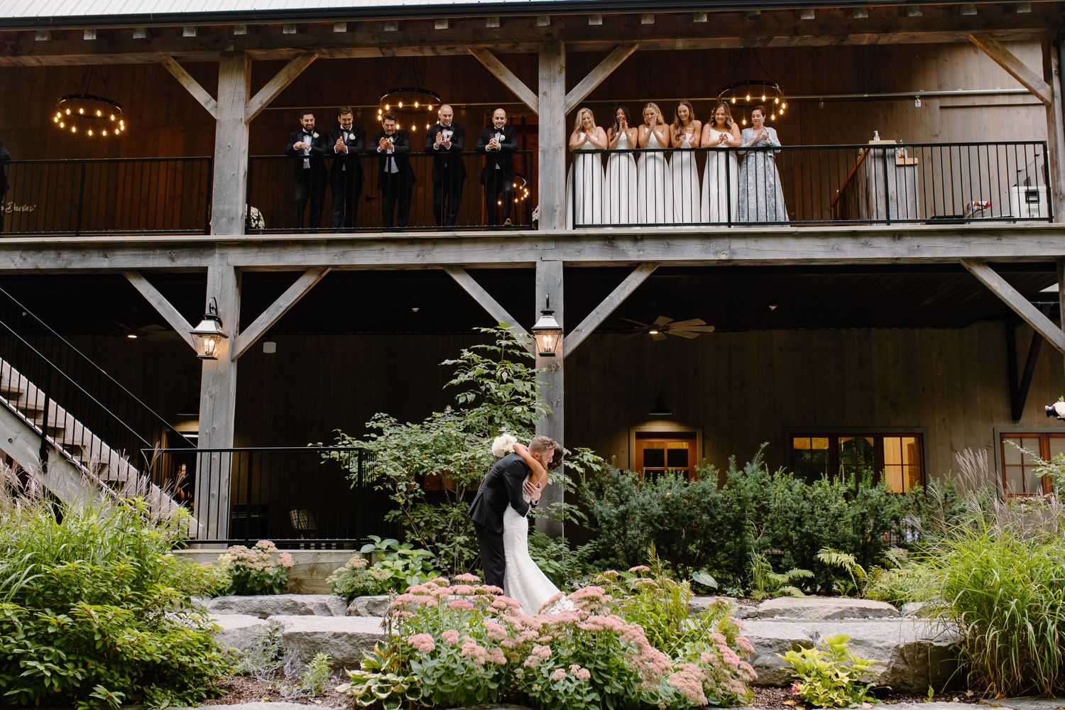 Wedding couple poses under rustic wooden beams with wedding party watching from balcony above.