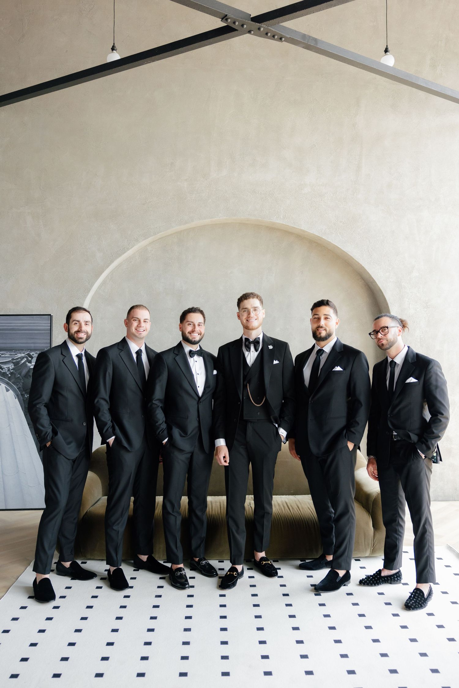 Wedding party group shot of groomsmen in dark suits posed against an arched wall.