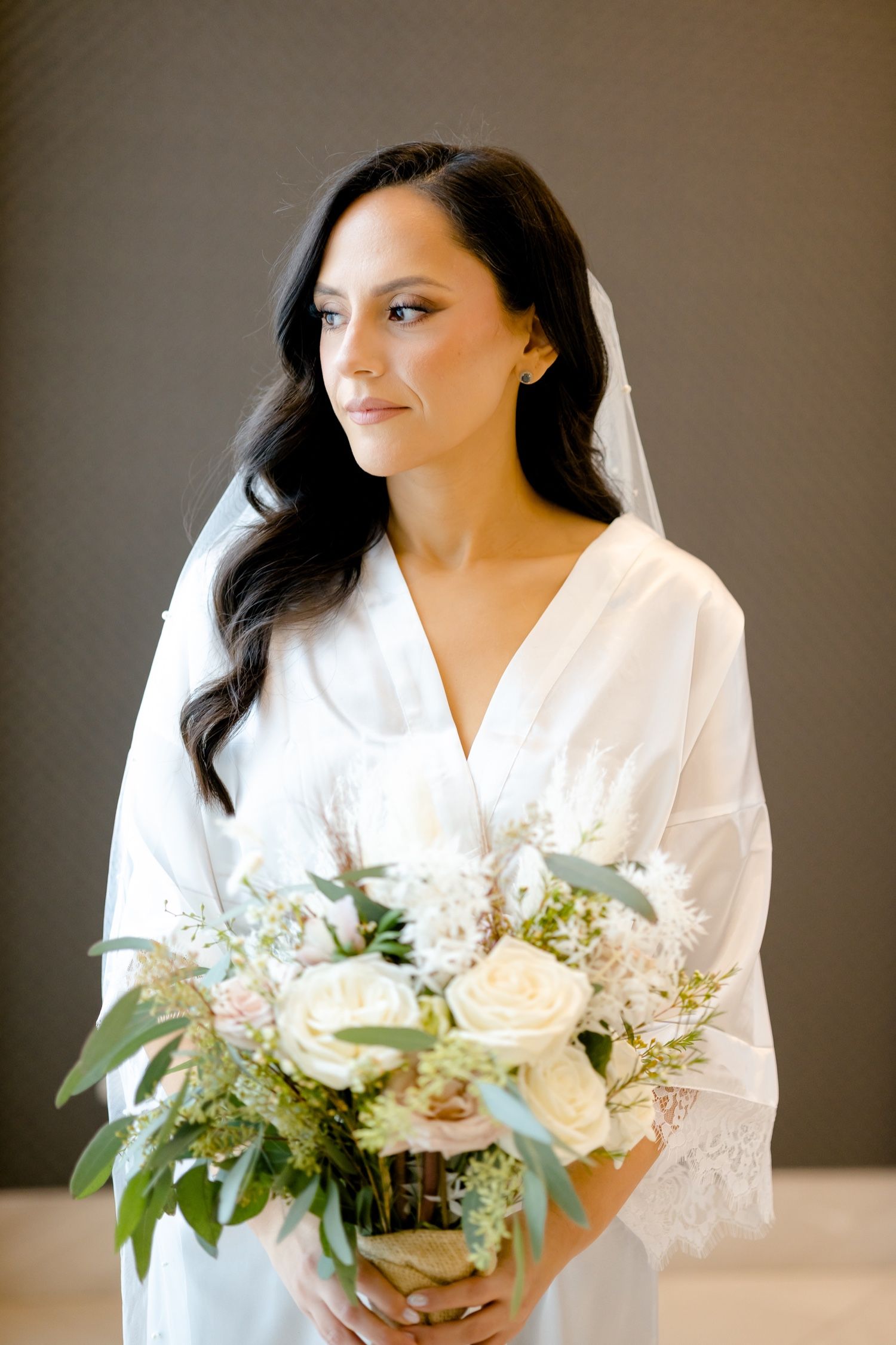 Bride in white robe holding white and green bouquet against grey background.