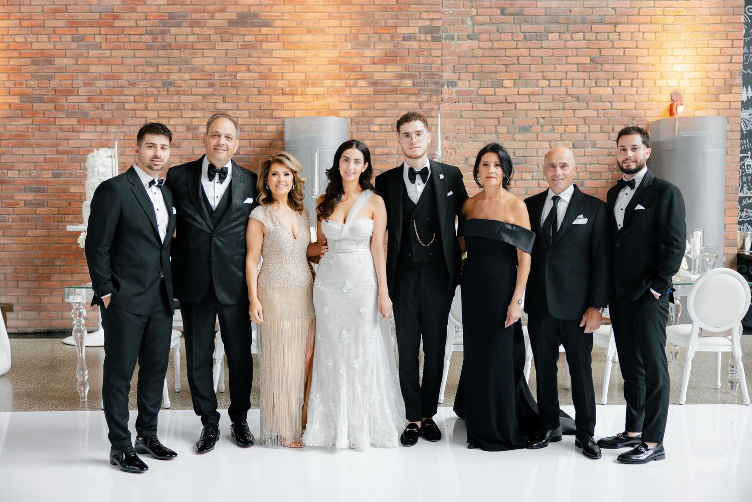 Wedding party group photo with eight people in formal attire against exposed brick wall.