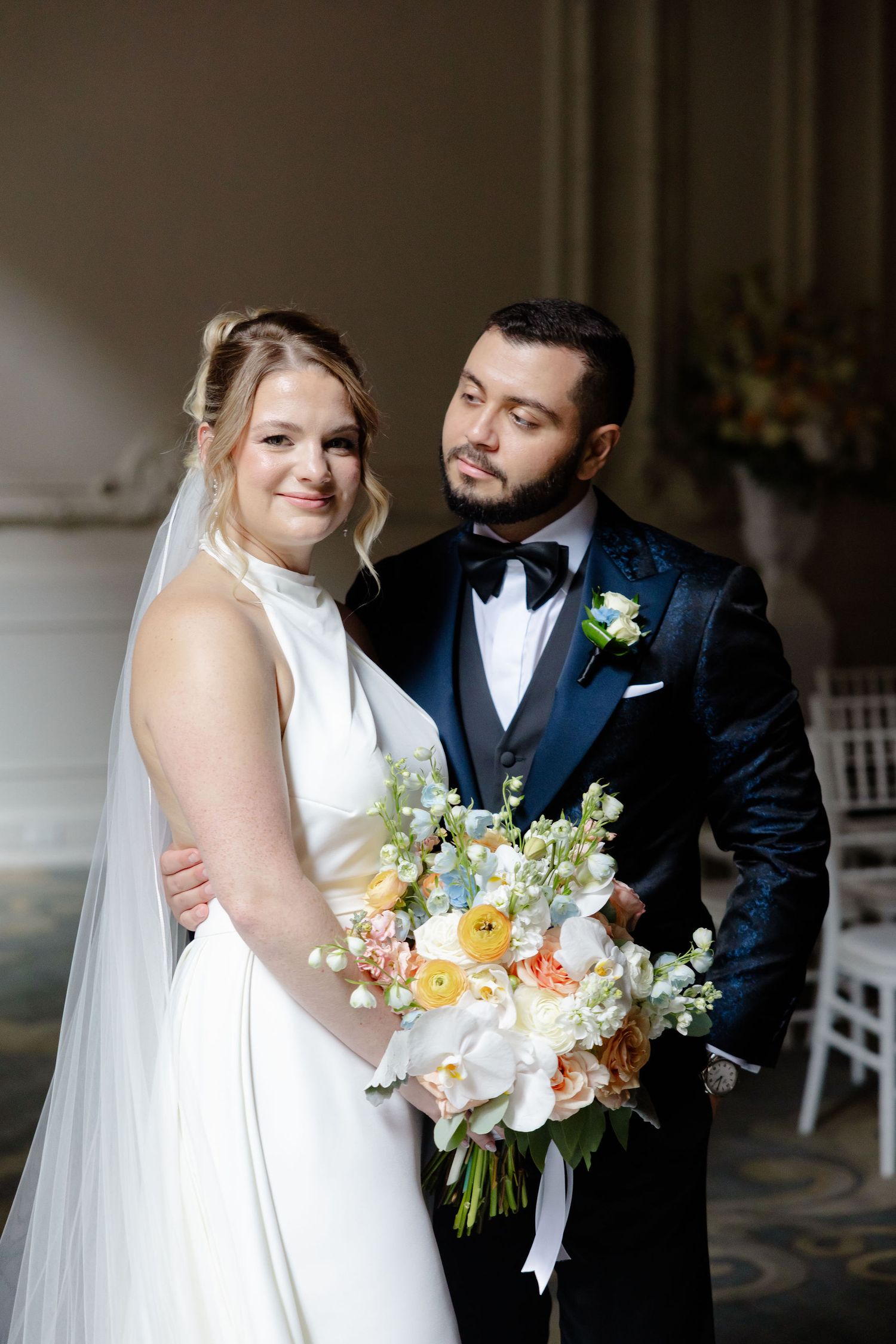 Elegant wedding couple poses together on a porch with a beautiful floral bouquet in soft pastel colors.