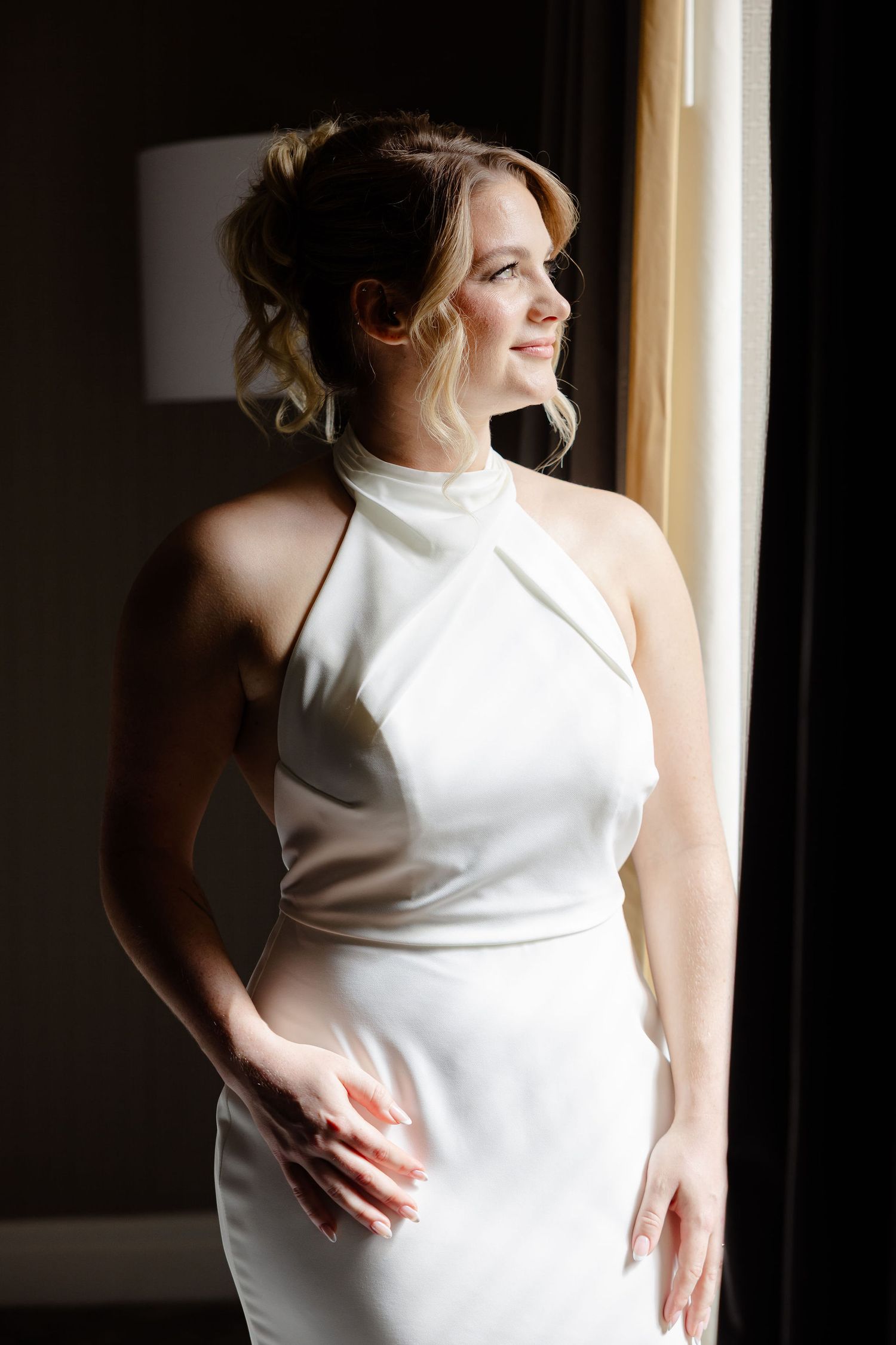 Elegant white halter wedding dress shown in dramatic window lighting.